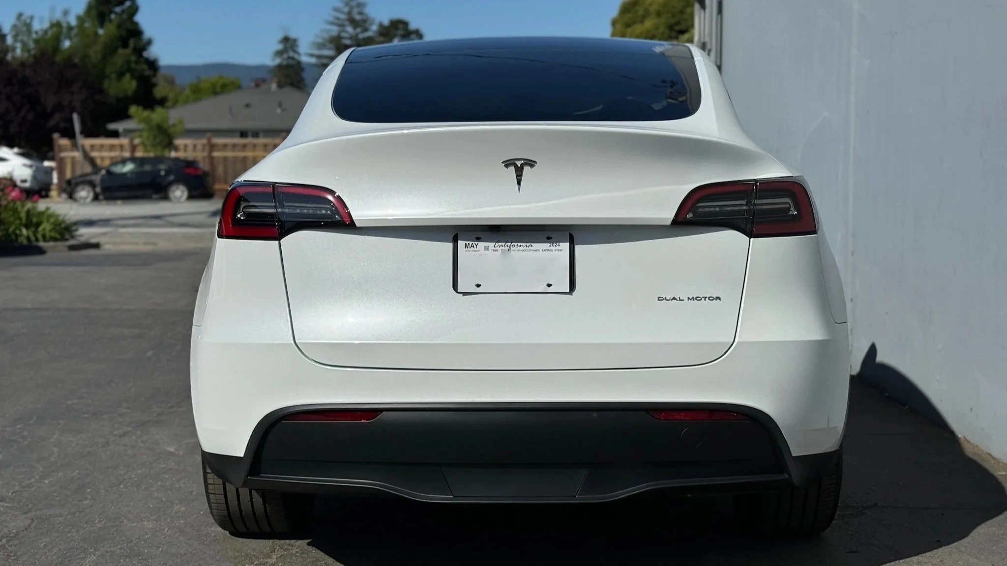 Rear view of a white Tesla Model Y electric vehicle parked next to a white wall, showing the Tesla logo, tail lights, license plate from California, and Dual Motor badge.