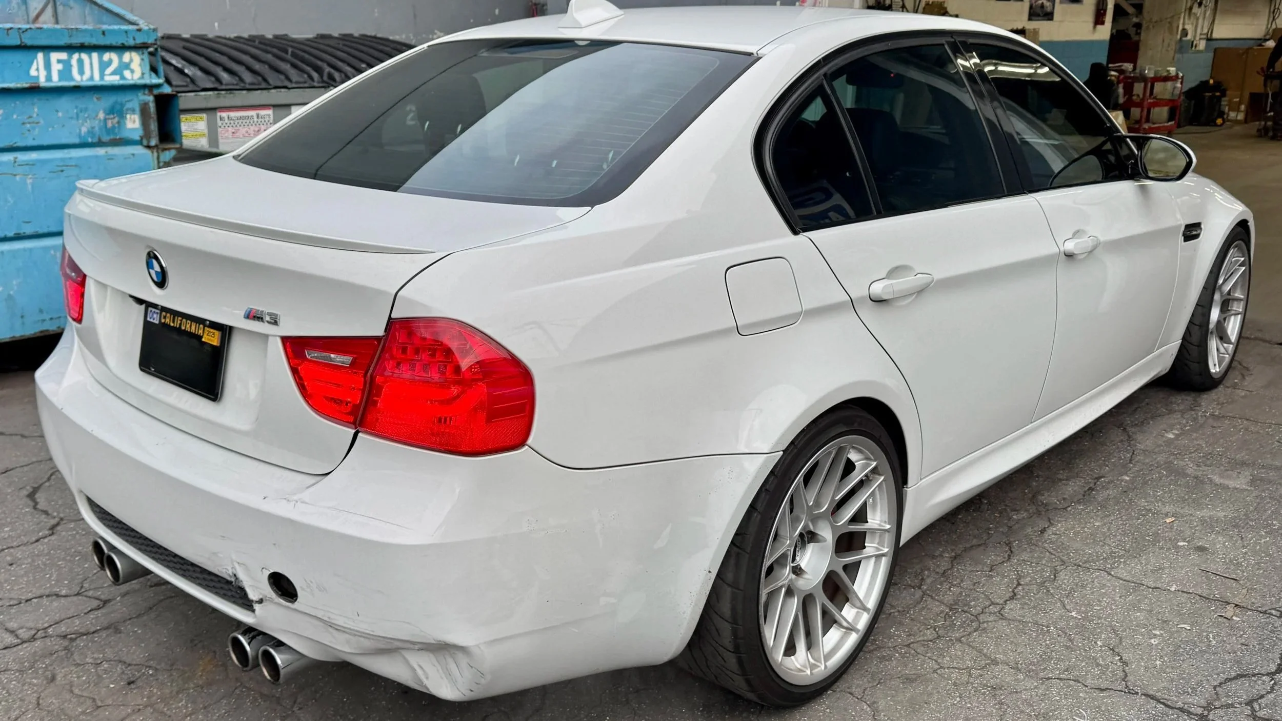 White BMW M3 sedan parked in an industrial area with cracked asphalt, showing rear and side view with event damage on the rear bumper.