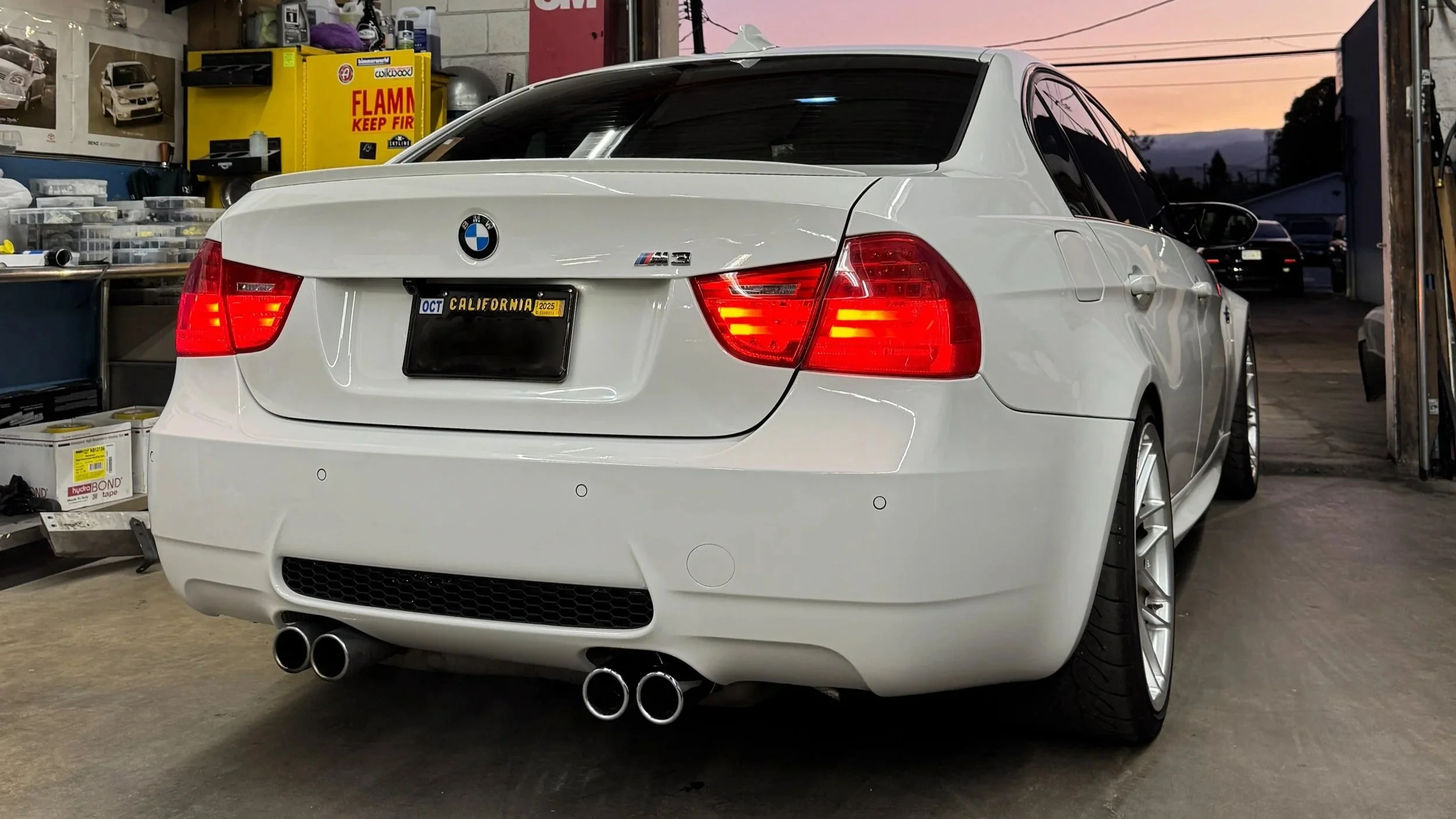 A white BMW M3 parked inside a garage with tools and supplies on shelves and a yellow flammable storage cabinet in the background. The car has a California license plate and quad exhaust pipes.