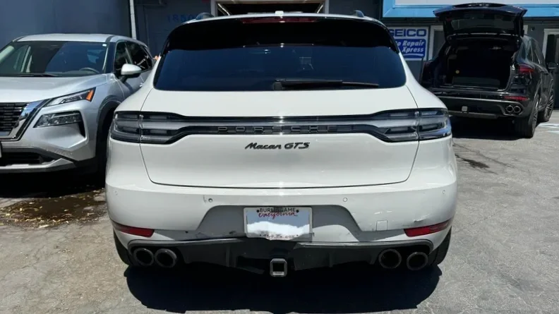 Rear view of a white Porsche Macan GTS SUV parked in a lot, with other vehicles nearby and an open trunk on a black SUV to the right.