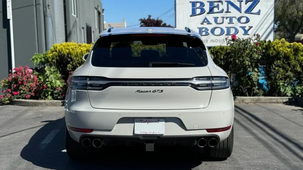 White Porsche Macan GTS parked in front of a Benz Auto Body shop, with pink and yellow flowers and trees in the background.