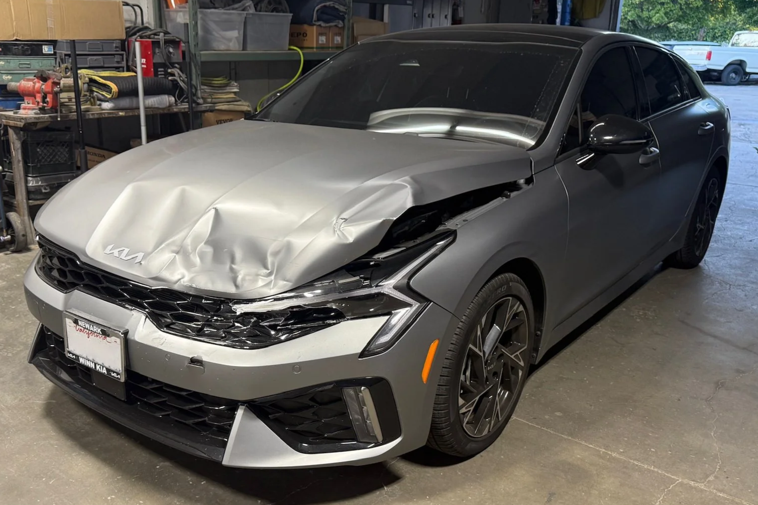 Gray Kia sedan with front end damage, crumpled hood, and missing front left headlight in a garage.