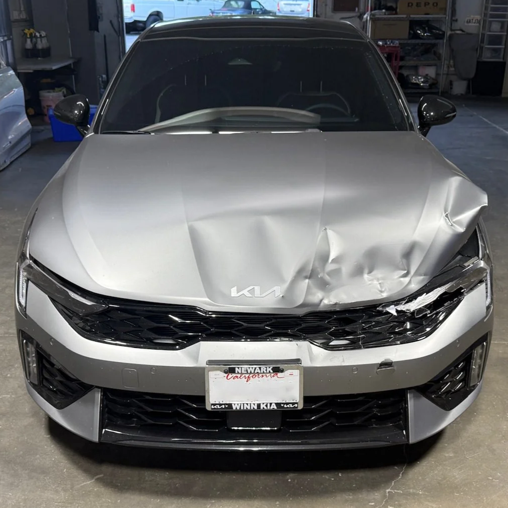 A silver Kia car with front-end damage, including a crumpled hood and a bent front fender, parked in a garage.