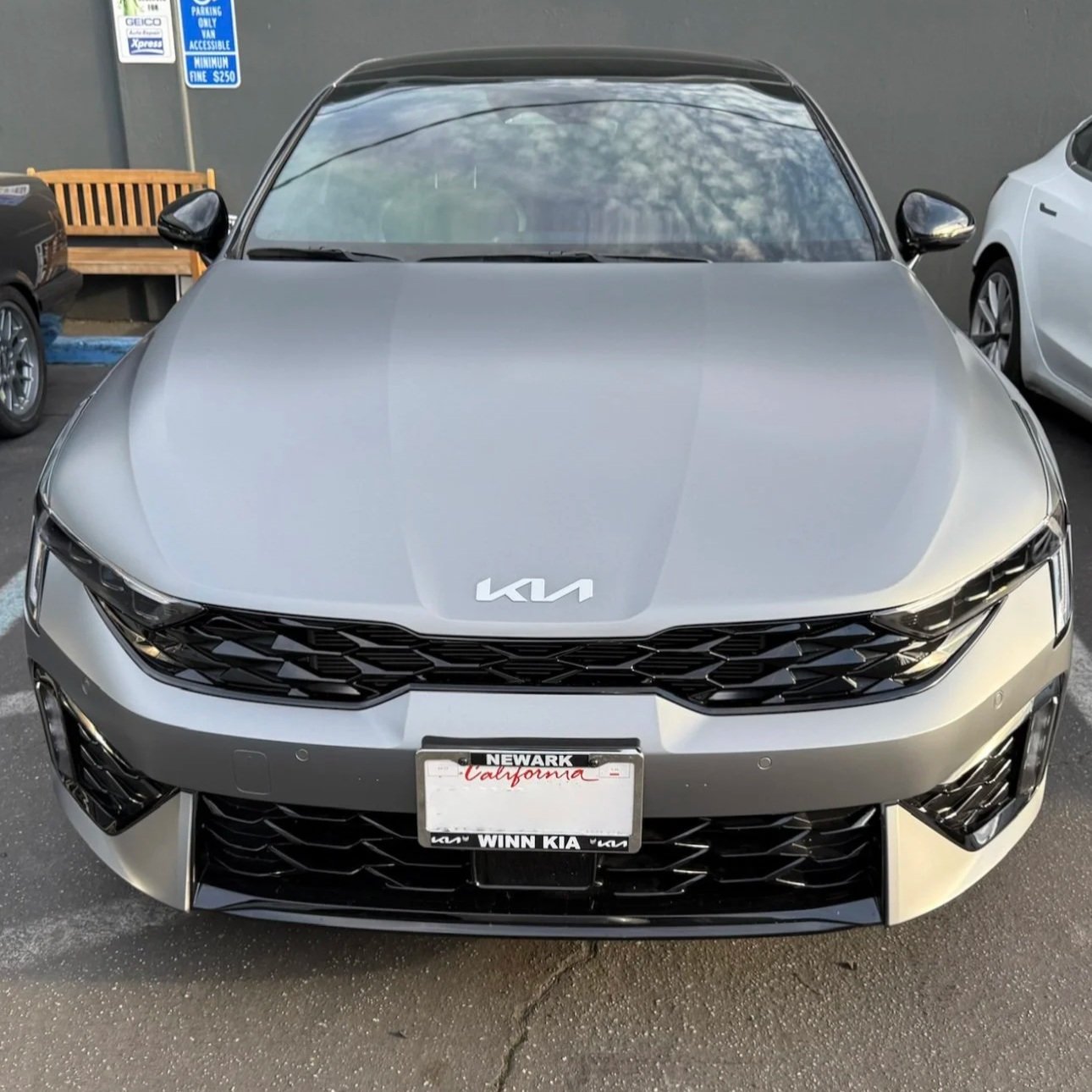 Front view of a silver Kia car parked in a parking lot, with a California license plate, next to a white car and a bench, with parking signs on a gray wall in the background.