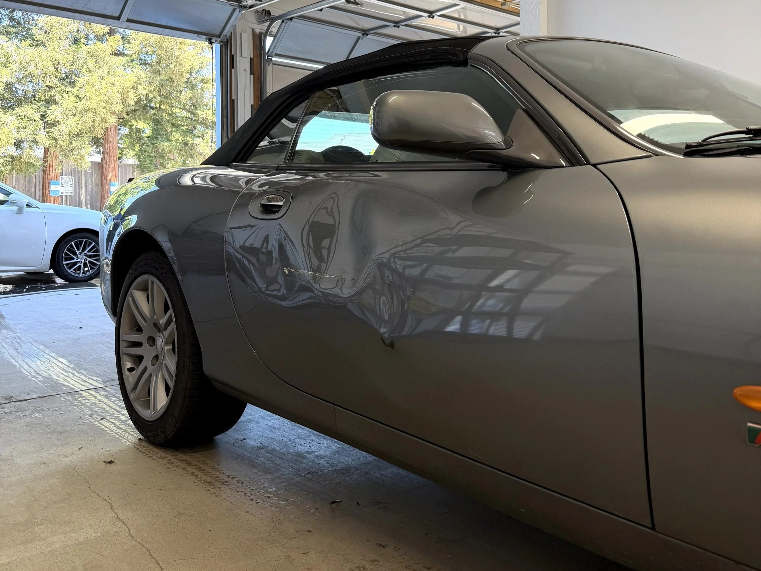 A silver convertible car parked inside a garage with an open door, with a white car visible in the background and trees visible outside.