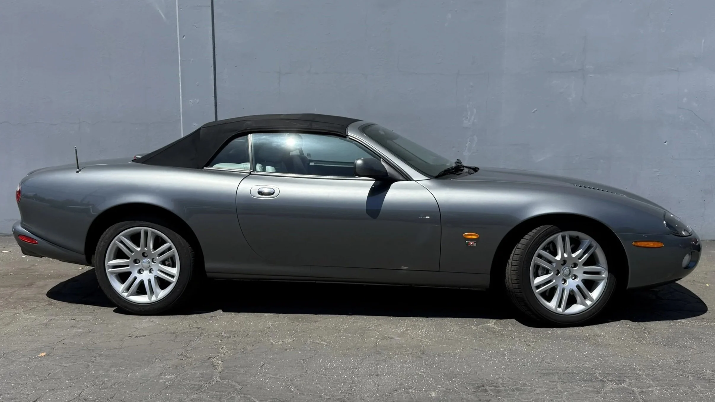 A silver convertible car with a black soft top parked against a gray wall.