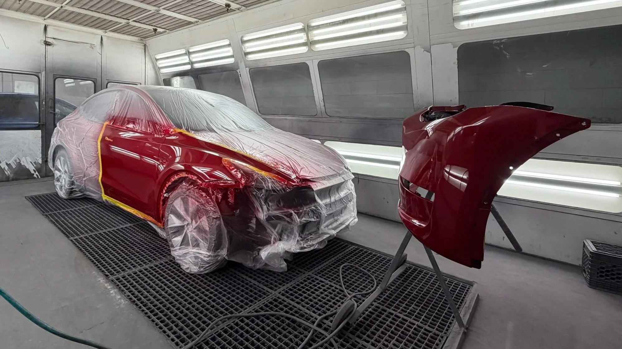 Car being painted in a spray booth, with a part of the car's body on a stand nearby, both covered in red paint.