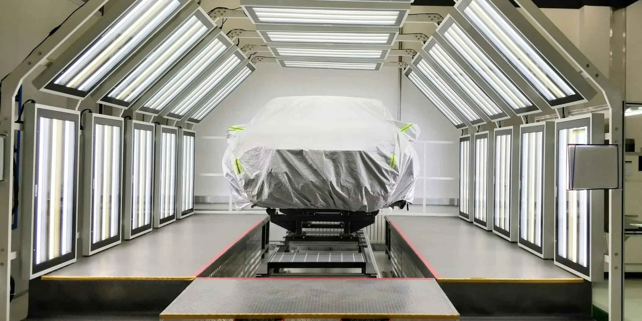 A car covered with a white protective sheet in a controlled environment with multiple ultraviolet lights.