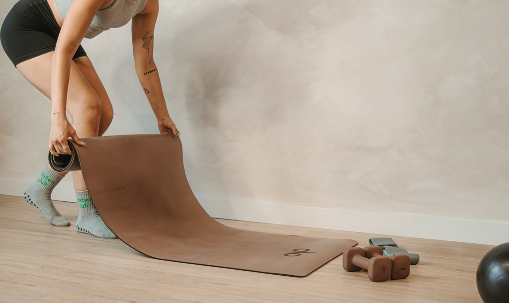 Person unrolling a Pilates mat beside hand weights while preparing for a workout session.