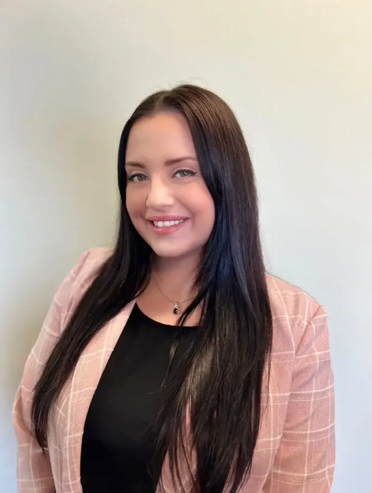 A young woman with long dark hair, smiling, wearing a pink plaid blazer and black top, standing against a plain light-colored wall.