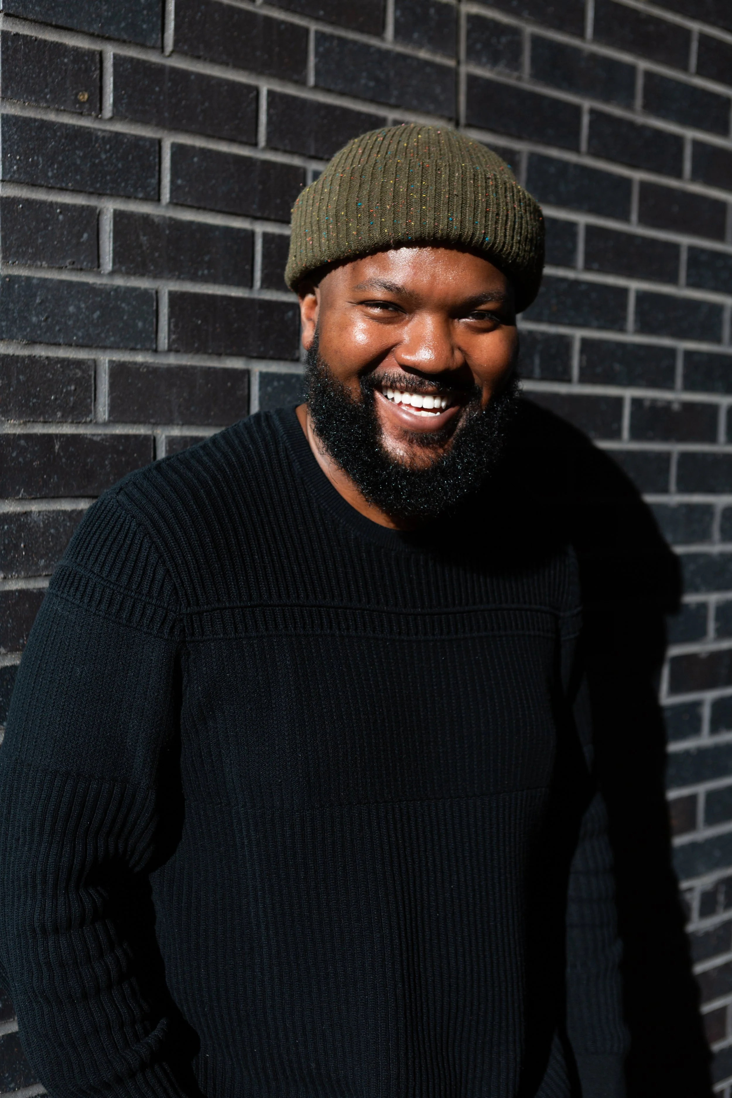 A smiling man with a beard wearing a green knit beanie and a black ribbed sweater, standing against a dark brick wall.