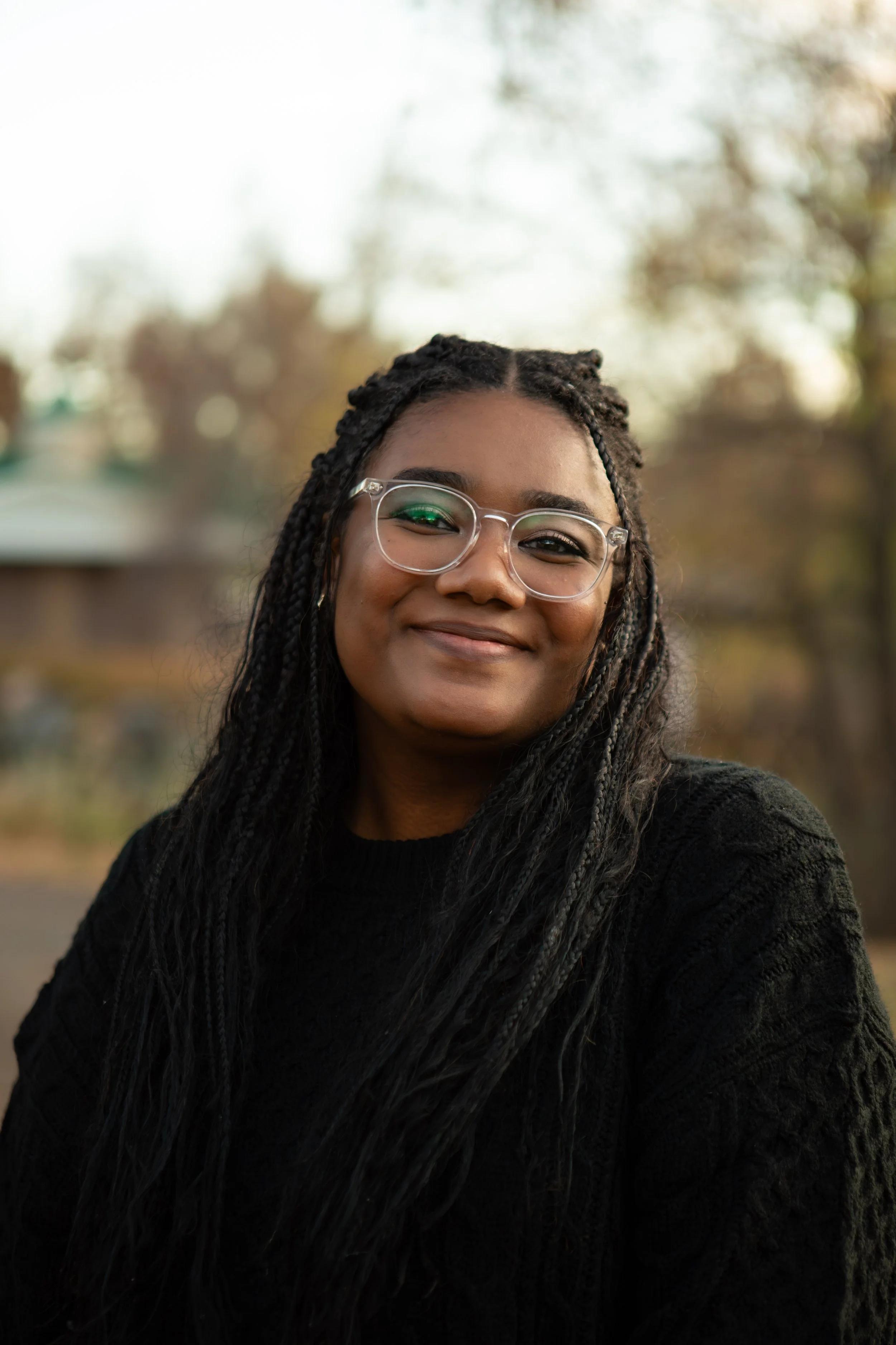 A young woman with glasses and braids smiling outdoors, with blurred trees in the background during autumn.