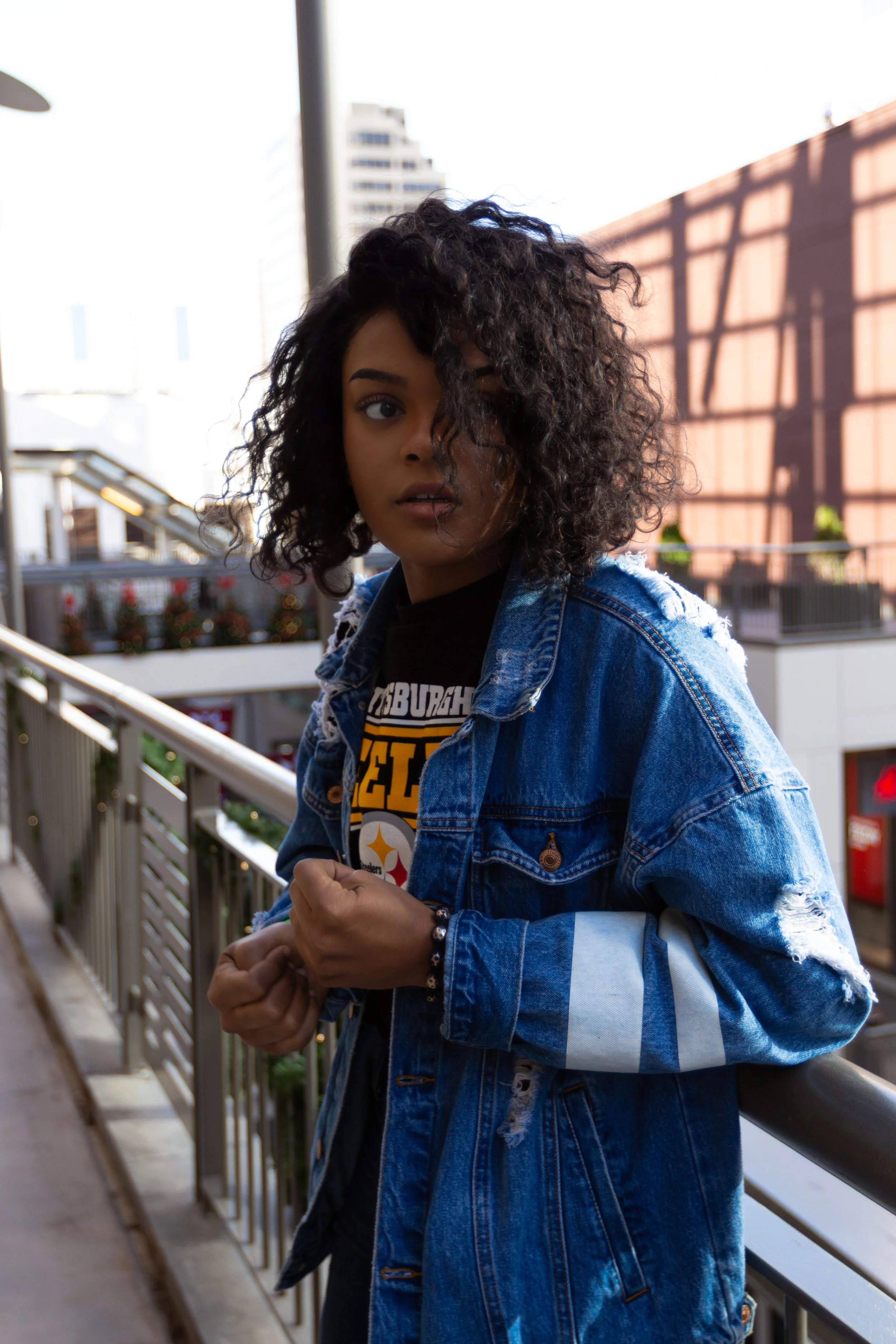 Young woman with curly hair wearing a denim jacket and a black Pittsburgh Steelers T-shirt, standing outdoors near a railing with a cityscape and buildings in the background.