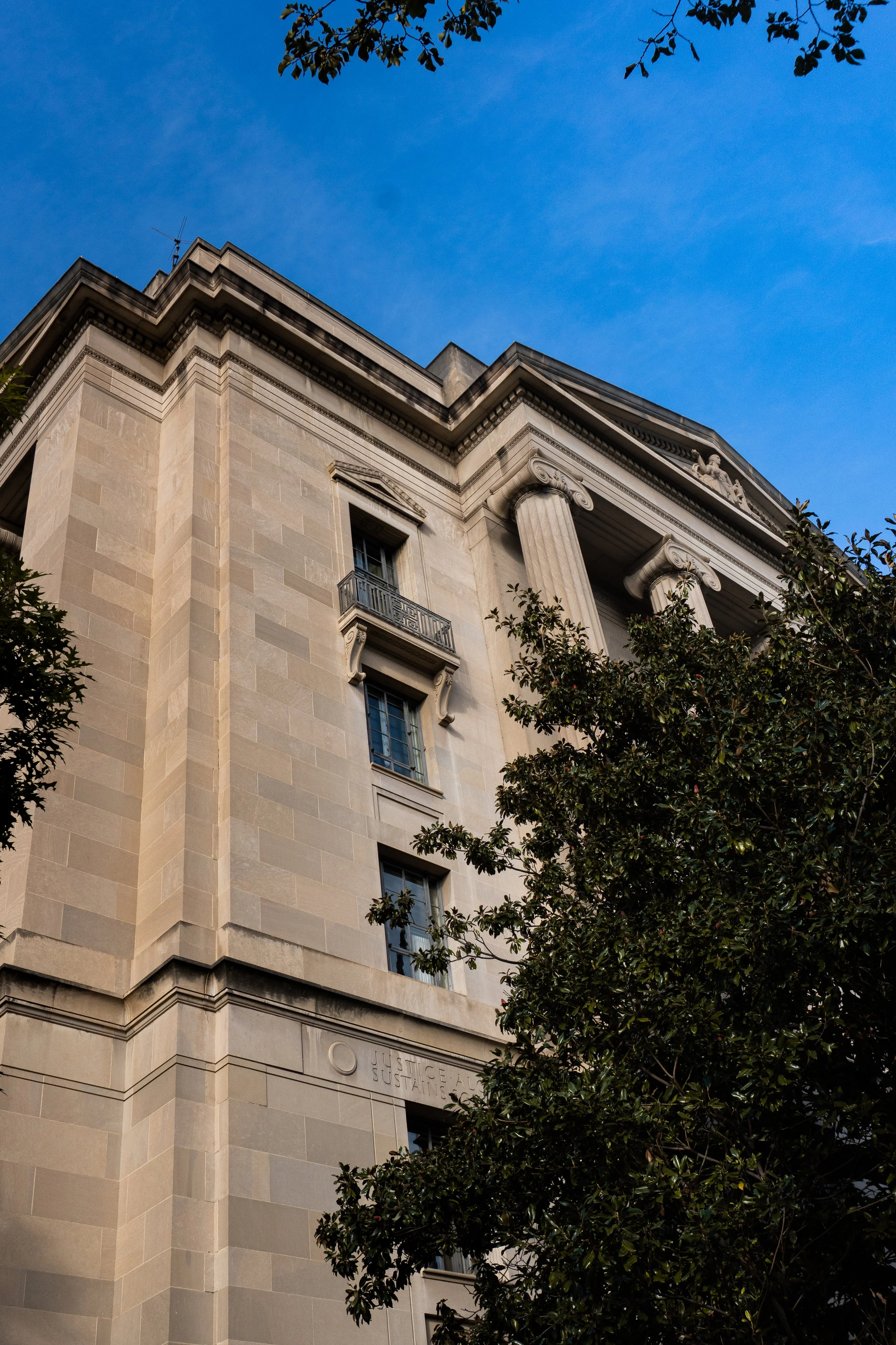 A close-up view of a classical-style building's upper facade showing columns and steps, partially obscured by tree branches with leaves, against a clear blue sky.
