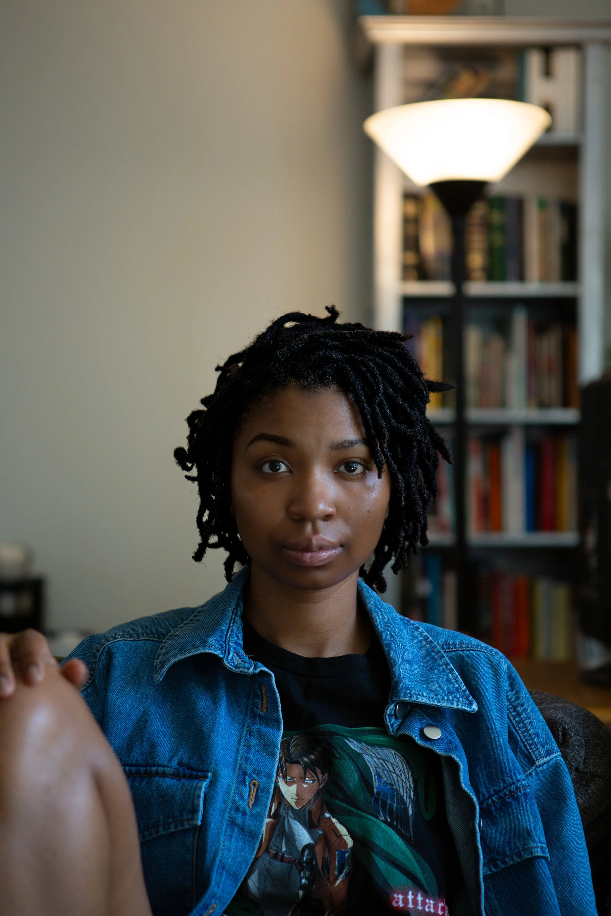 A young woman with dreadlocks and medium skin tone sitting indoors, wearing a denim jacket over a graphic T-shirt, with a bookshelf and a lamp in the background.