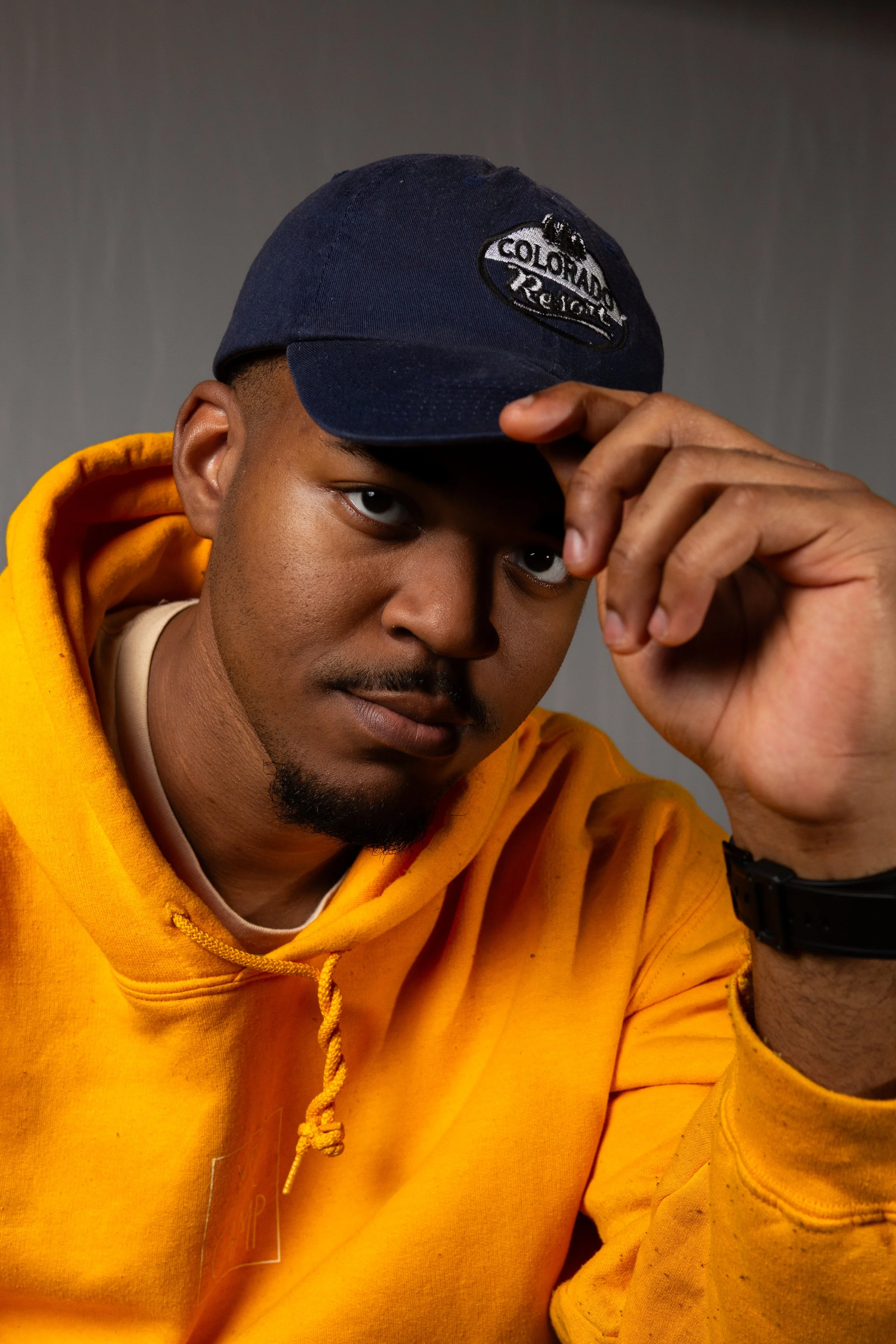 A young man wearing a yellow hoodie and a navy blue cap, adjusting the cap while looking at the camera. The background is plain and gray.