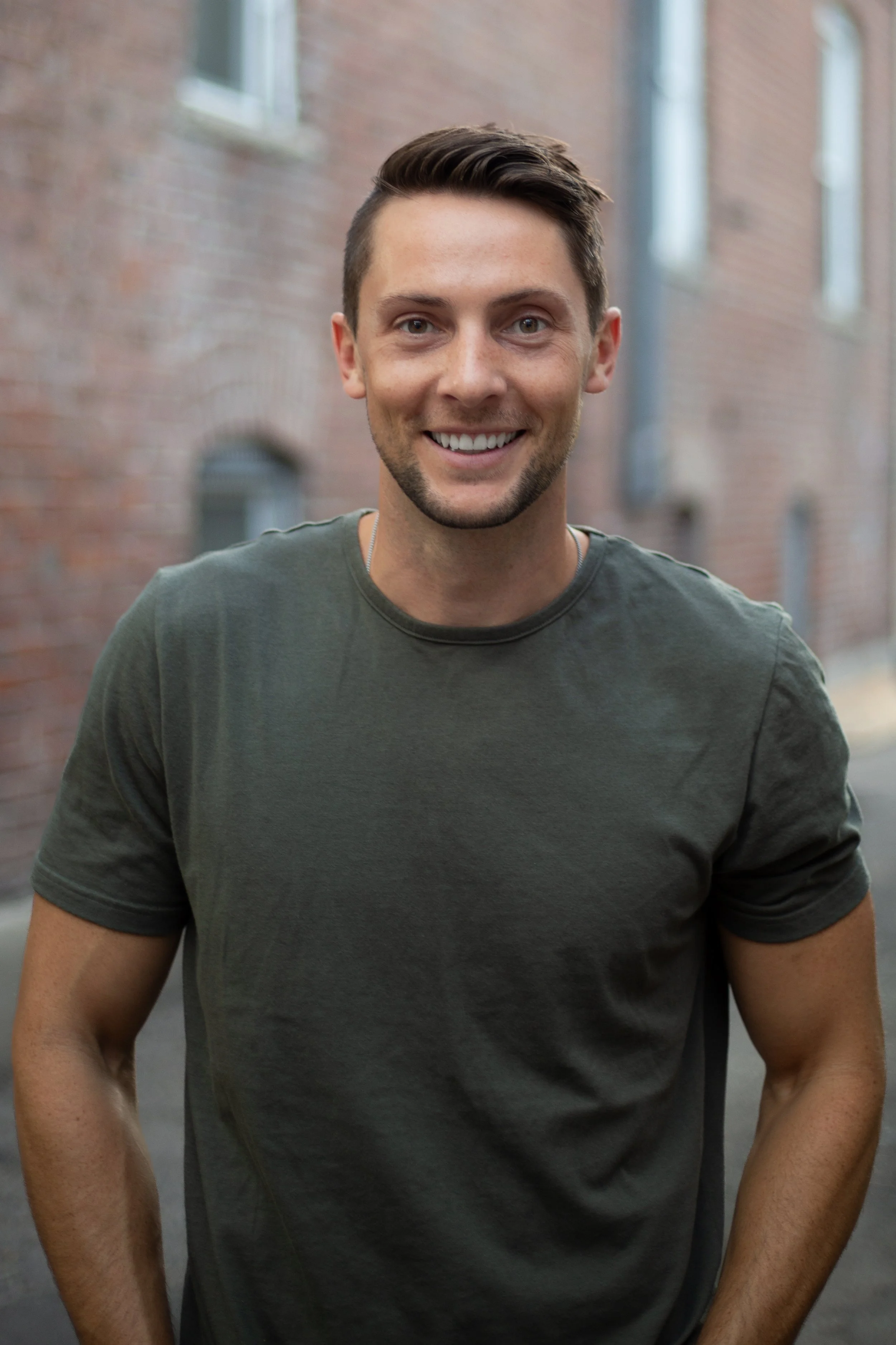 A young man with short brown hair, blue eyes, and a light beard smiling outdoors against a blurred background of brick buildings.