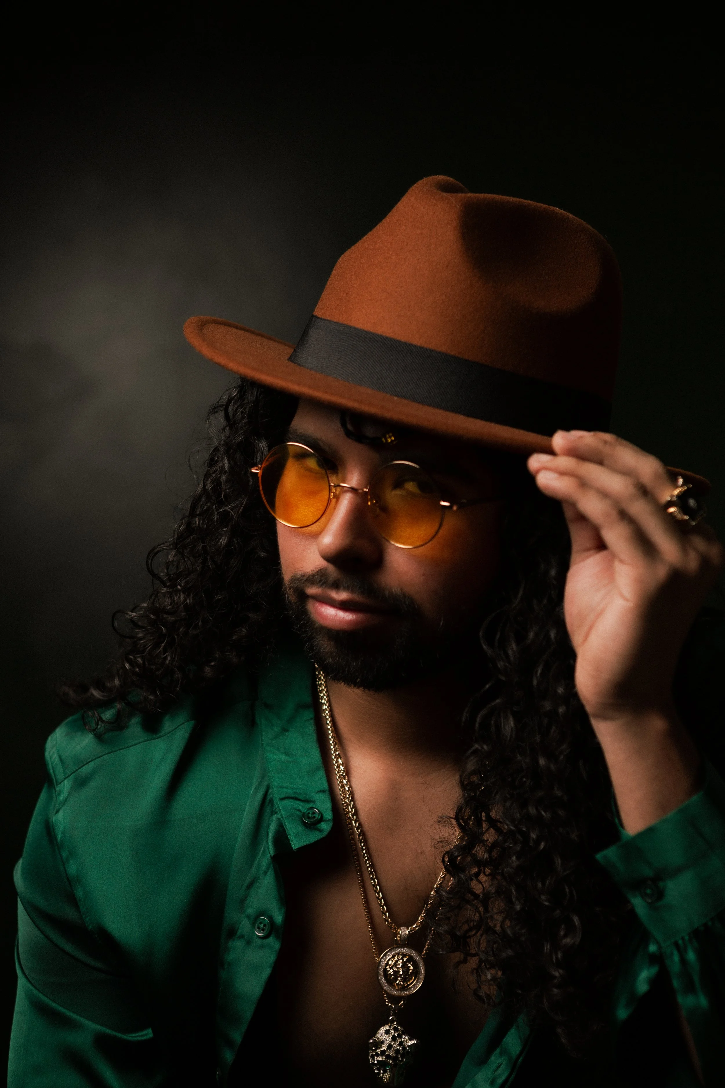 A man with curly hair and a beard wears a brown hat, yellow-tinted sunglasses, a green button-up shirt, and gold jewelry against a dark background.