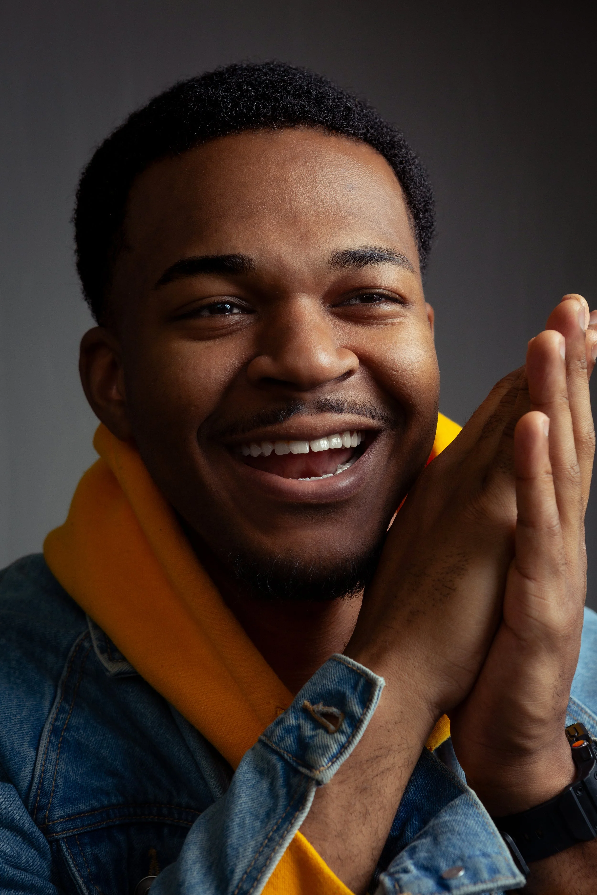 Close-up of a smiling man with dark skin, wearing a denim jacket and a yellow hoodie, posed with hands together near his face against a dark background.