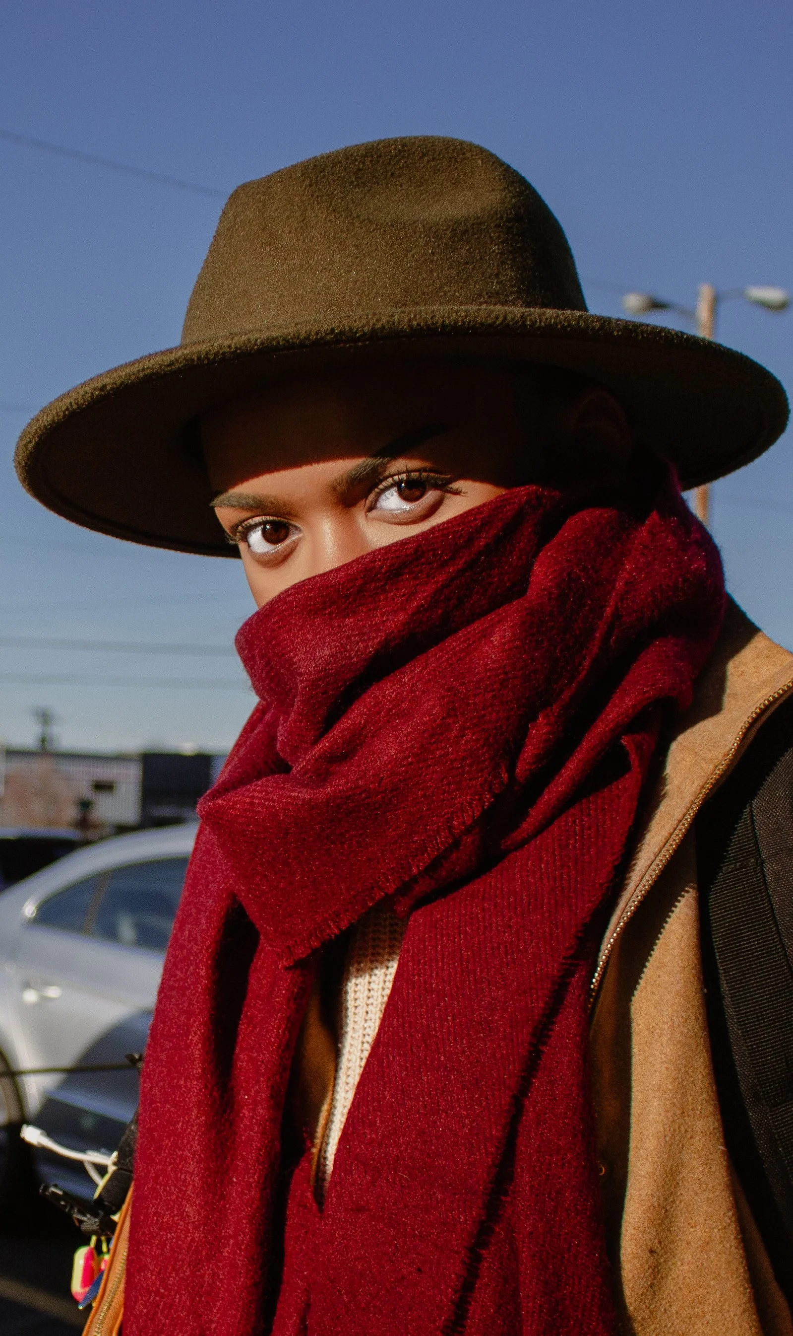 Person wearing a wide-brimmed hat, a red scarf covering their face, and a beige jacket, standing outdoors in daylight with a clear blue sky and a parking lot in the background.