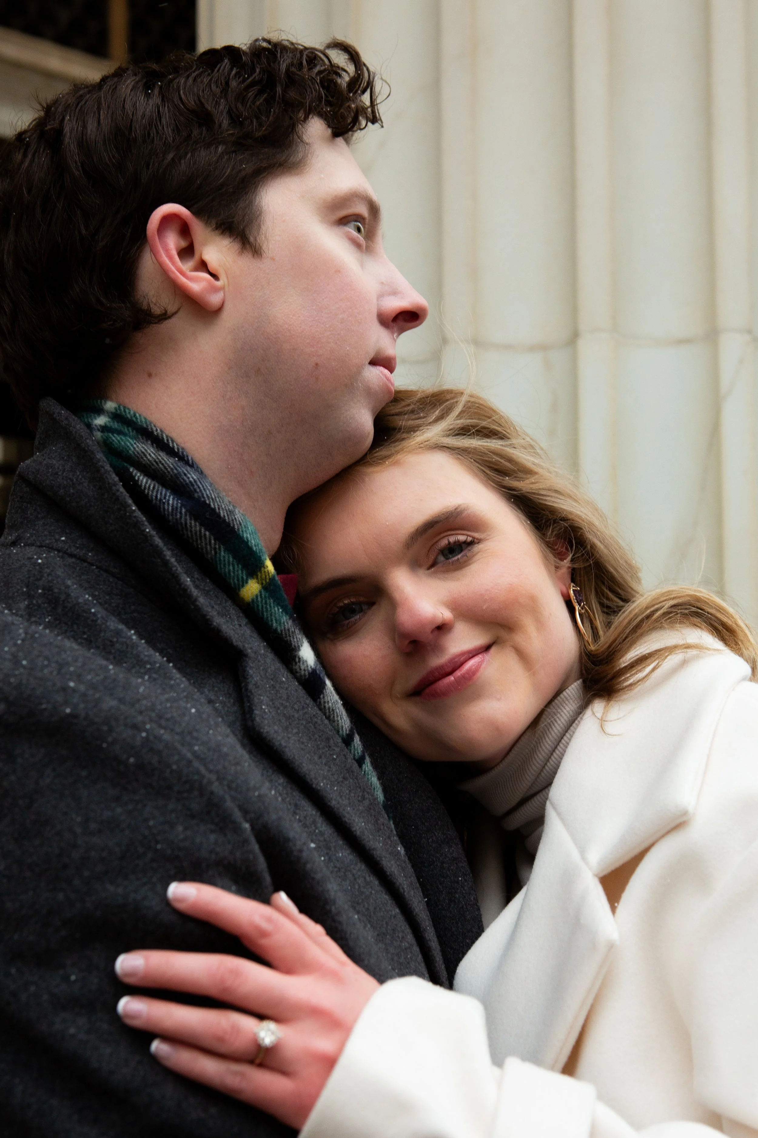 A man with short, curly brown hair and light skin, and a woman with blonde hair and light skin, embracing with the woman's head resting on the man's chest, both smiling softly.