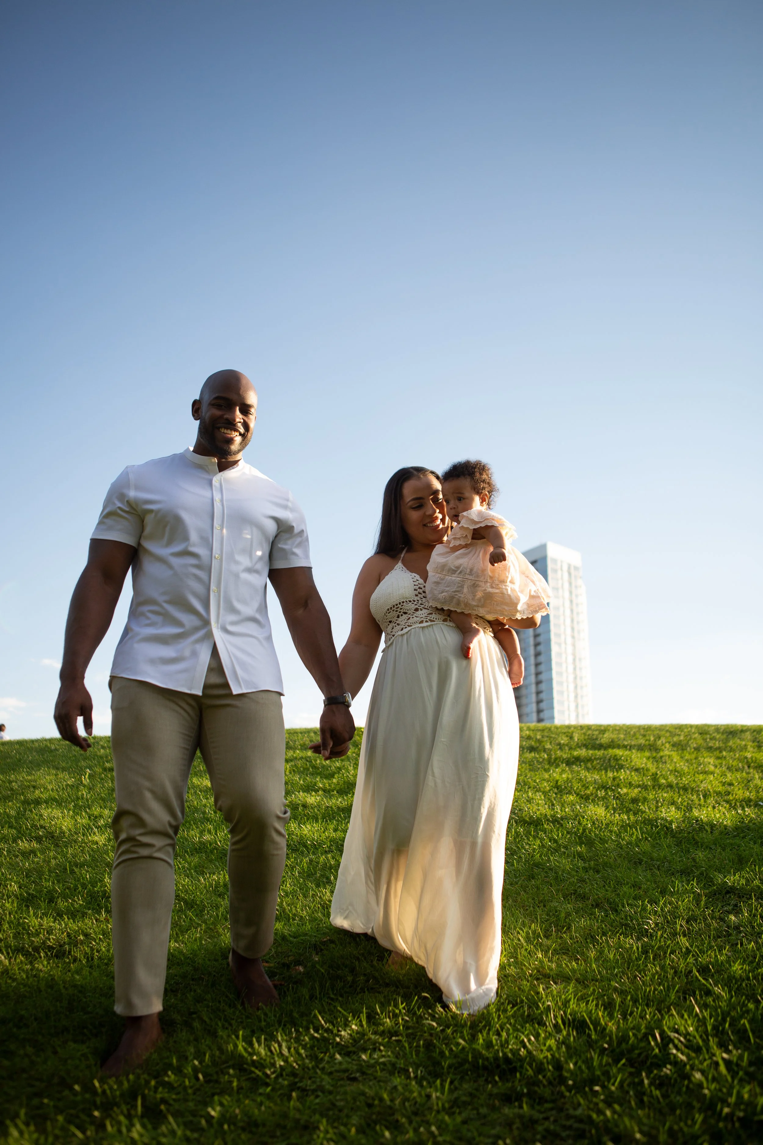 A family of three walking in a park under a clear blue sky, with a tall building in the background. The man is wearing a white shirt and beige pants, smiling. The woman is holding a baby girl dressed in a light-colored dress, and she is smiling as they walk.