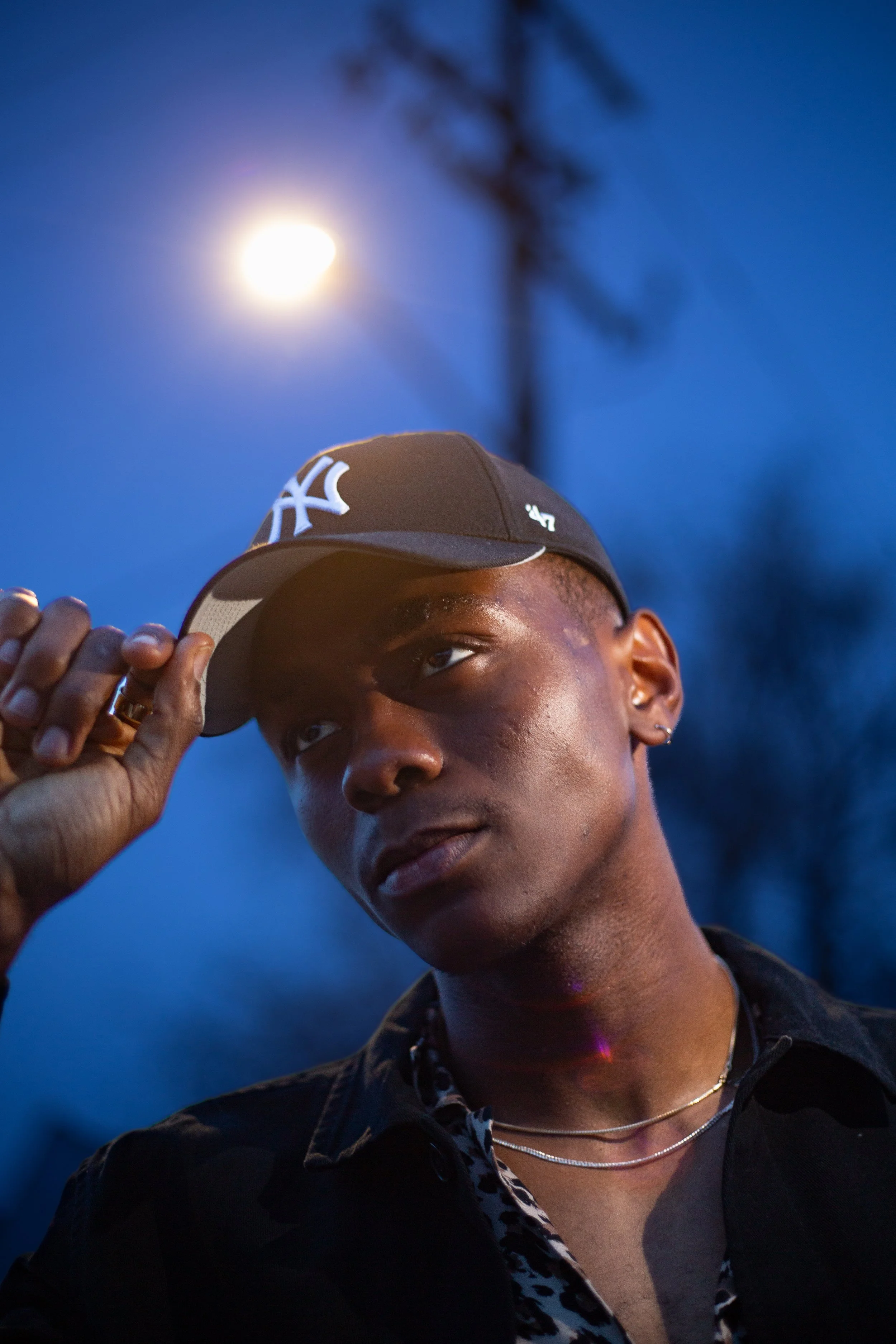 A young person adjusts their cap with a New York Yankees logo during dusk, with the moon and a blurred tree in the background.
