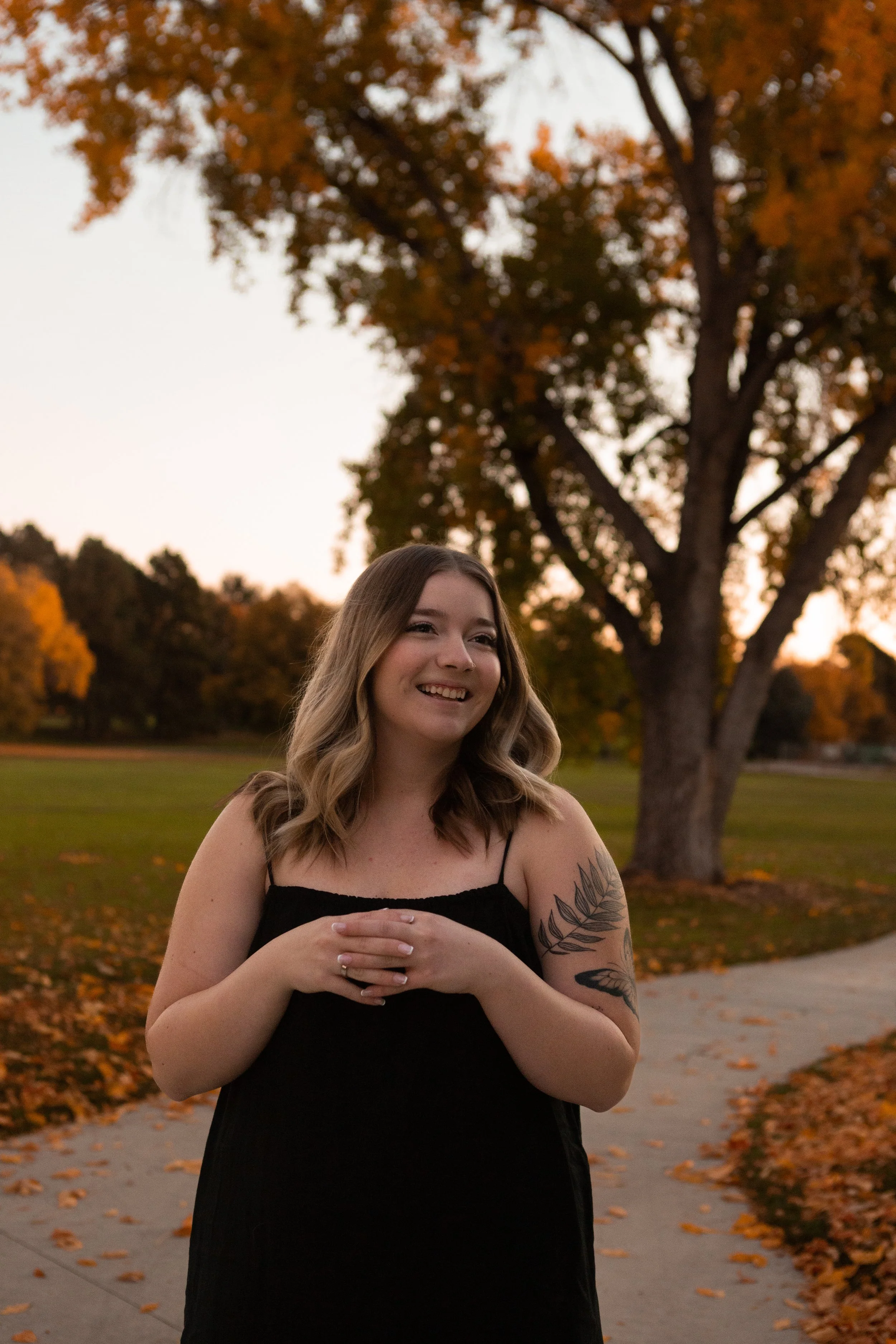 A smiling woman with wavy blonde hair wearing a black spaghetti strap dress, standing outdoors during fall with leafless trees and fallen orange leaves on the ground.