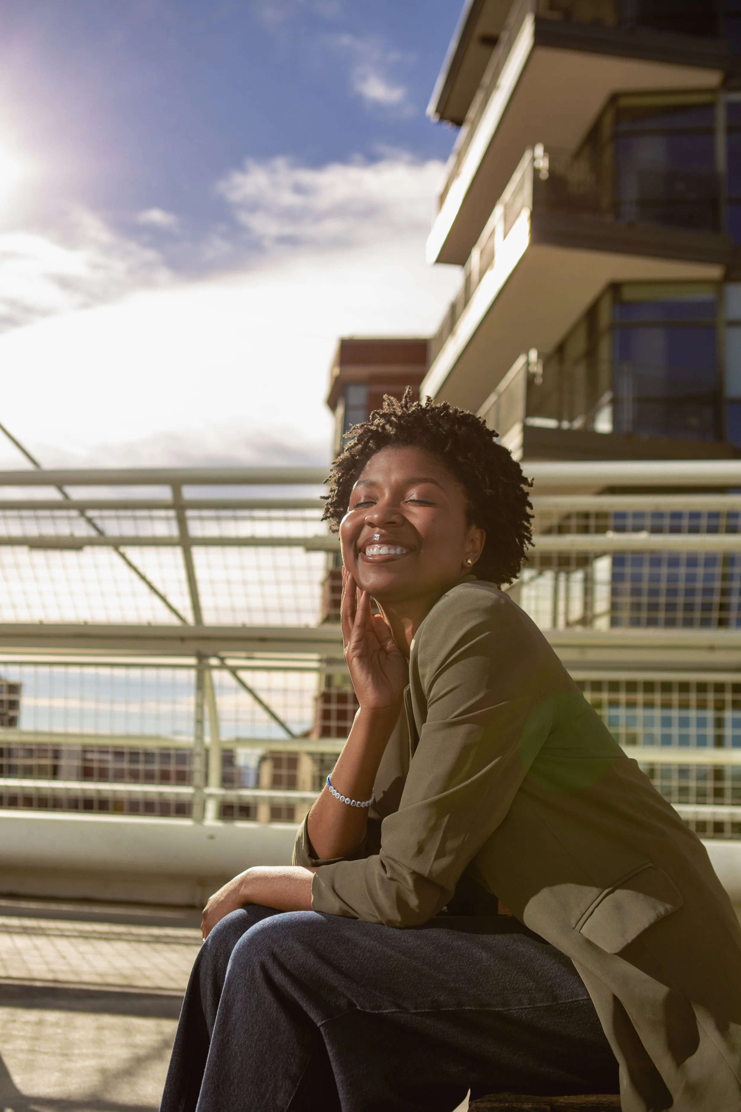 A woman with short curly hair smiling, sitting on a bench outdoors under a partly cloudy sky, with modern buildings and balconies in the background.