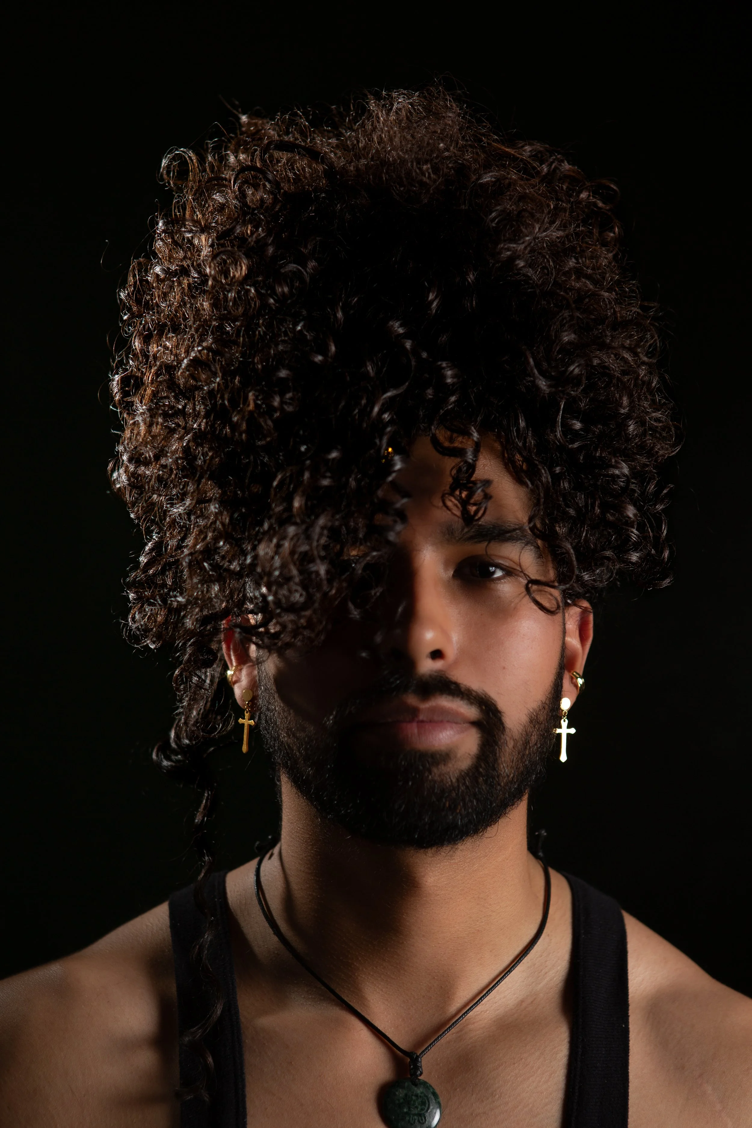A person with curly hair styled in a tall, voluminous manner, wearing earrings with crosses, a black tank top, and necklaces, against a black background.