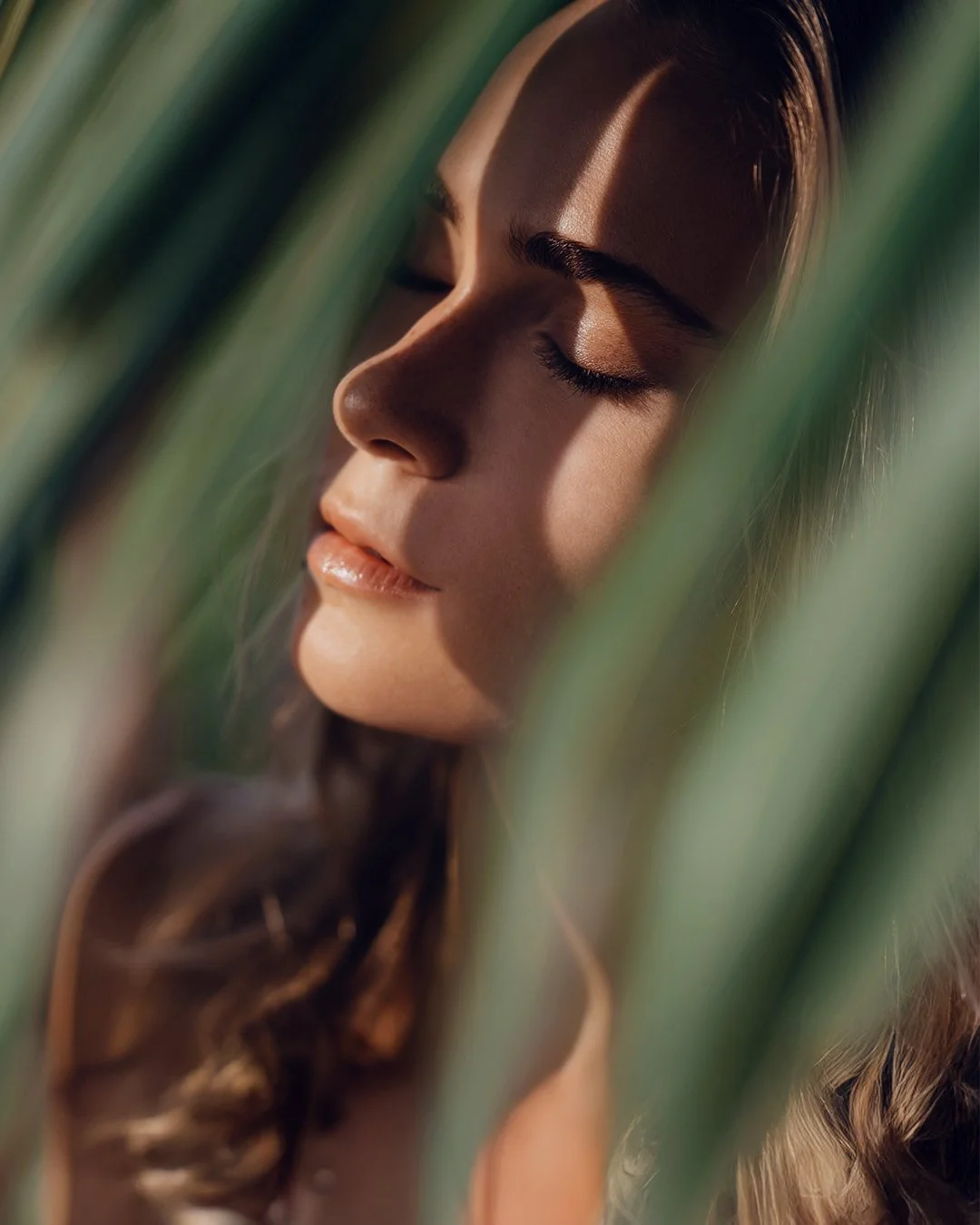 A close-up of a woman with her eyes closed, surrounded by green leaves, with sunlight casting a shadow on her face.