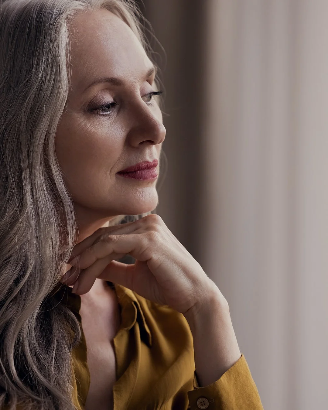 A woman with long, wavy hair and light makeup, wearing a mustard-colored blouse, resting her chin on her hand and looking thoughtfully out of a window.