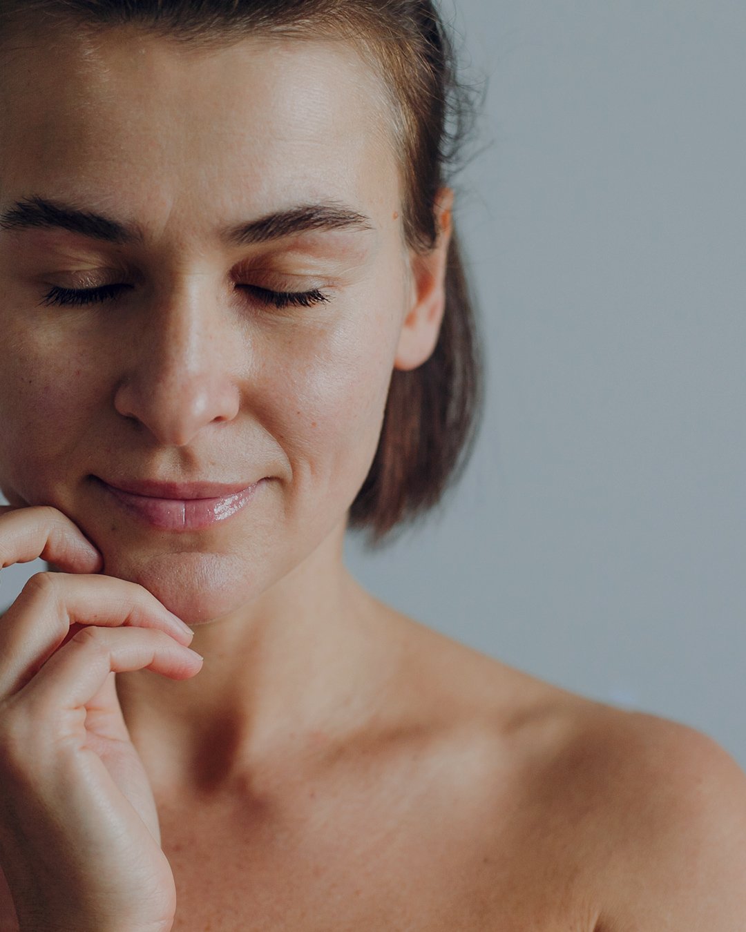 Close-up of a woman with closed eyes and a gentle smile, touching her chin with her hand, against a plain gray background.