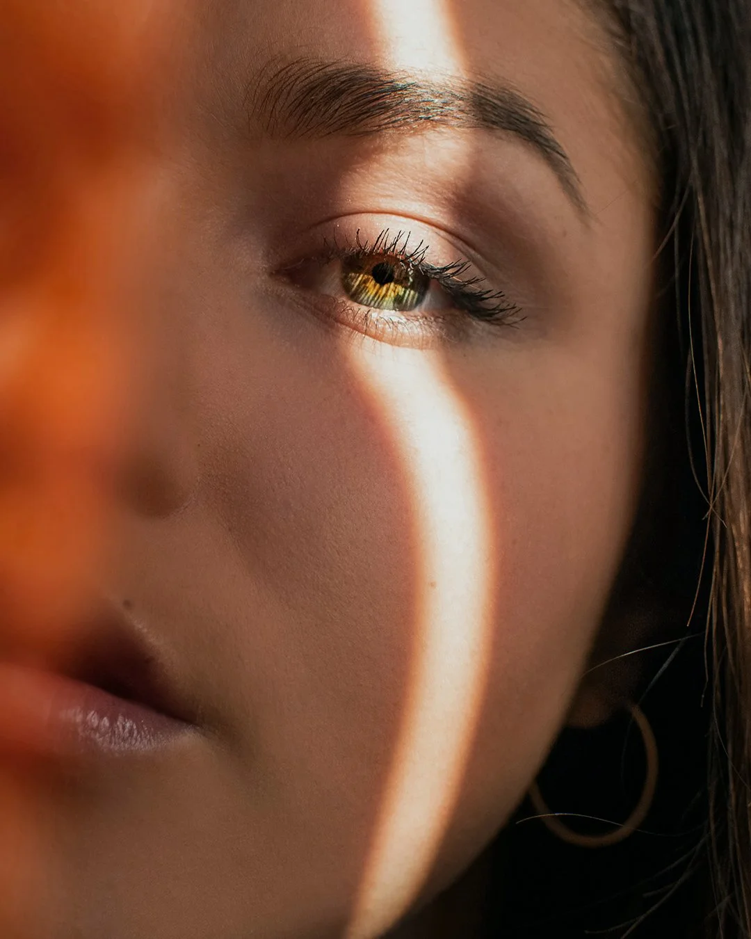 Close-up of a woman's face showing her eye, cheek, and part of her lips, with sunlight casting a bright streak across her skin.