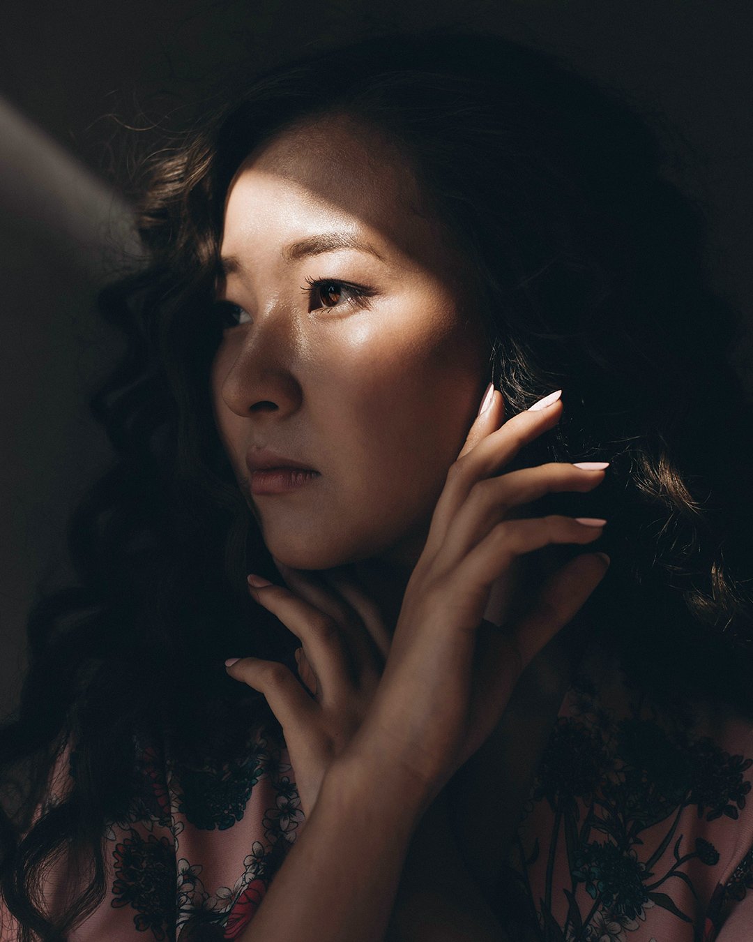 A woman with wavy dark hair gazing thoughtfully, illuminated by a soft light highlighting her face, with her hand gently touching her cheek.