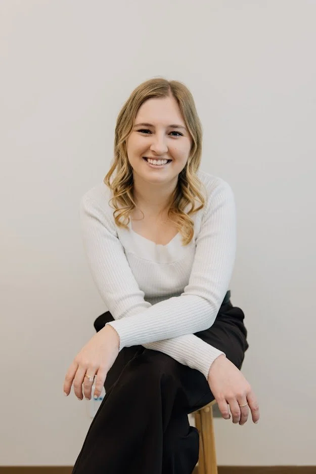 A young woman with blonde hair sitting on a stool against a plain light gray wall, smiling at the camera.