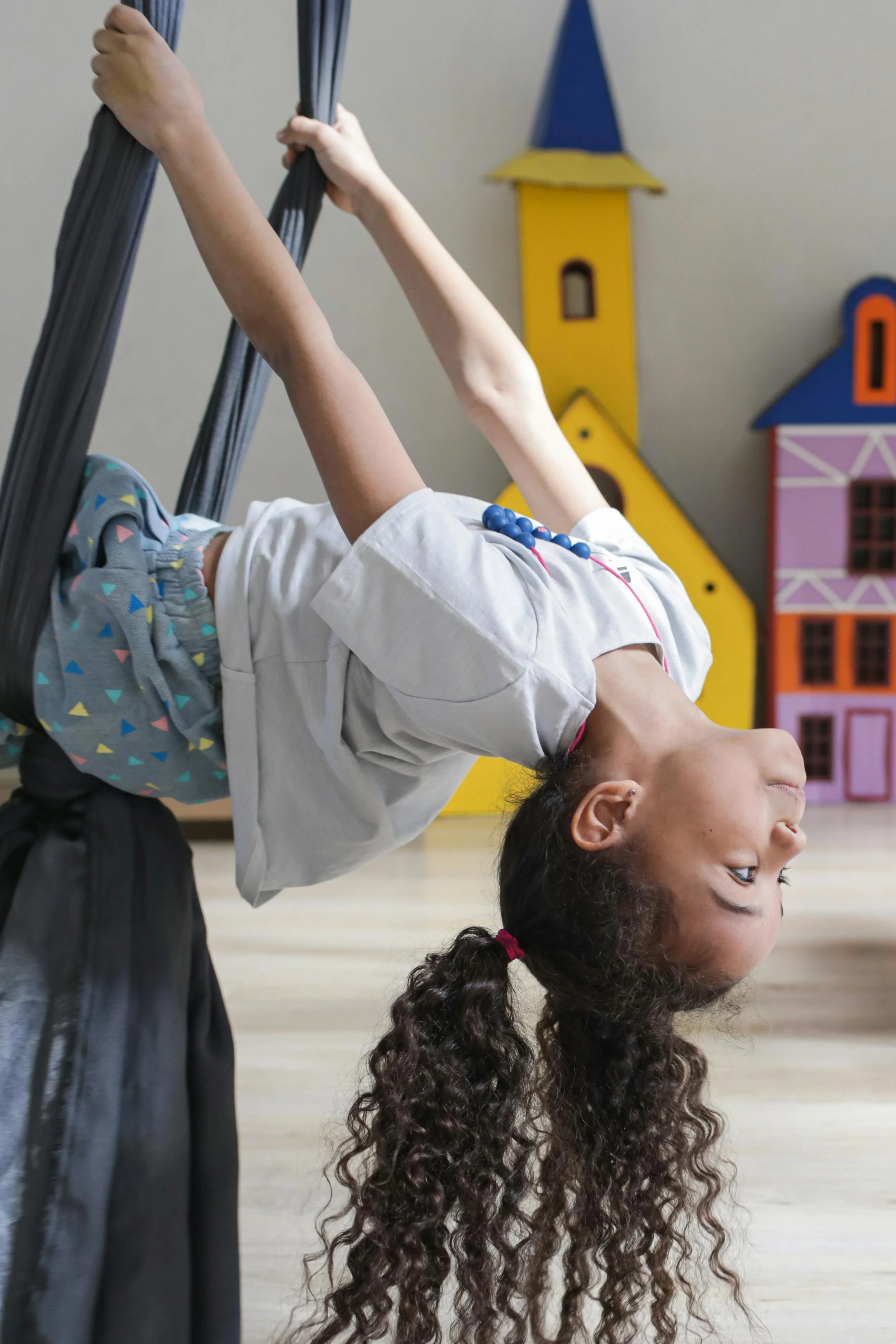 A young girl with curly brown hair tied in pigtails hanging upside down from a black gymnastic ring, with a background of colorful wooden toy houses.