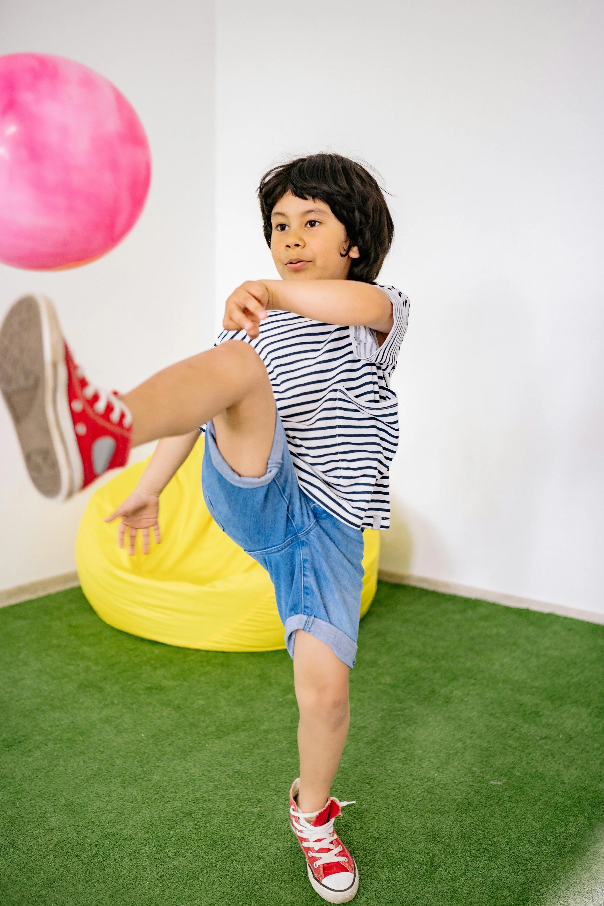 Child playing indoor with a pink ball, wearing a striped shirt, denim shorts, and red sneakers.