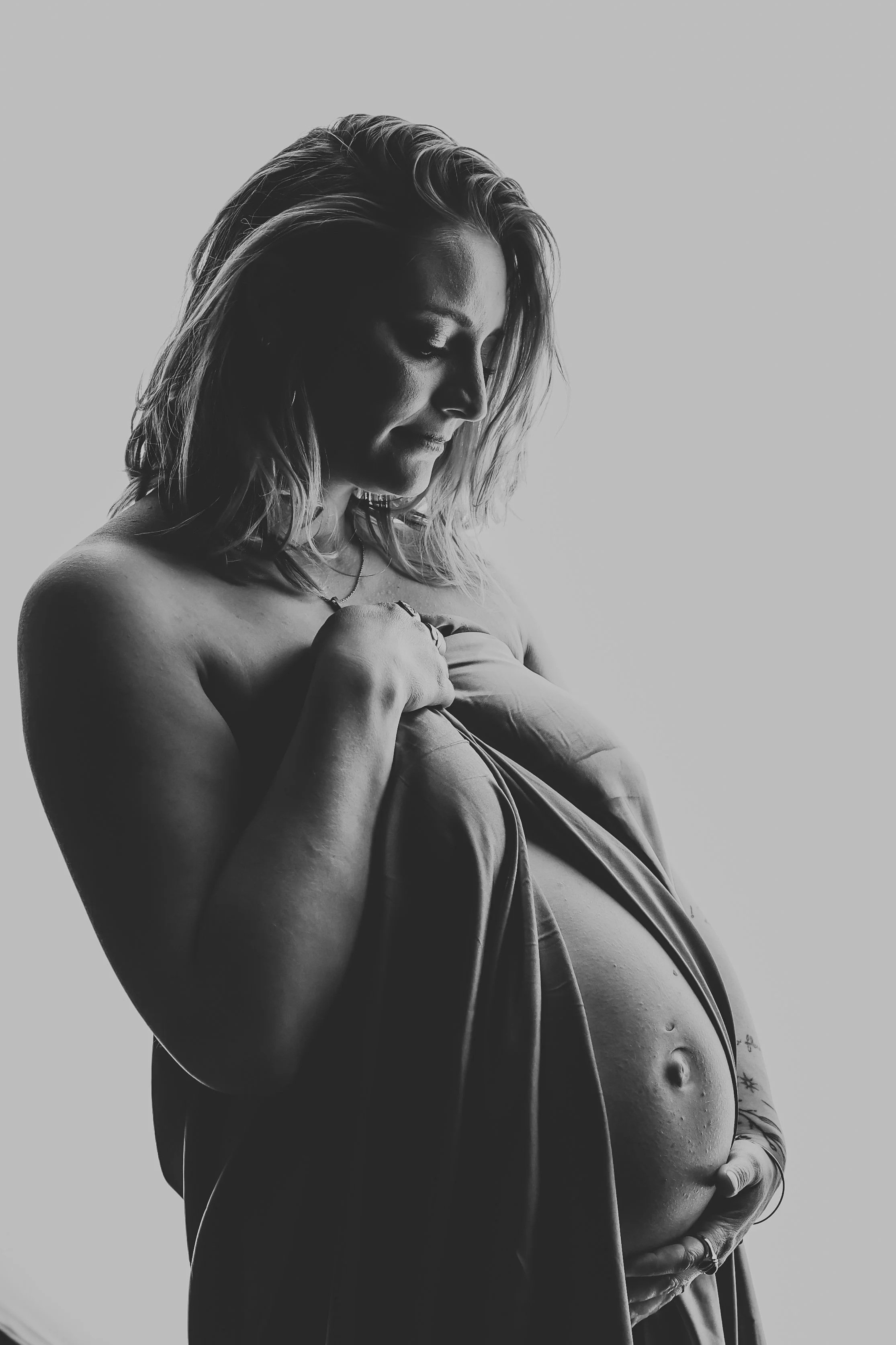 Black and white photo of a pregnant woman looking down, holding her baby bump with both hands, with a serene expression, and her hair slightly tousled.