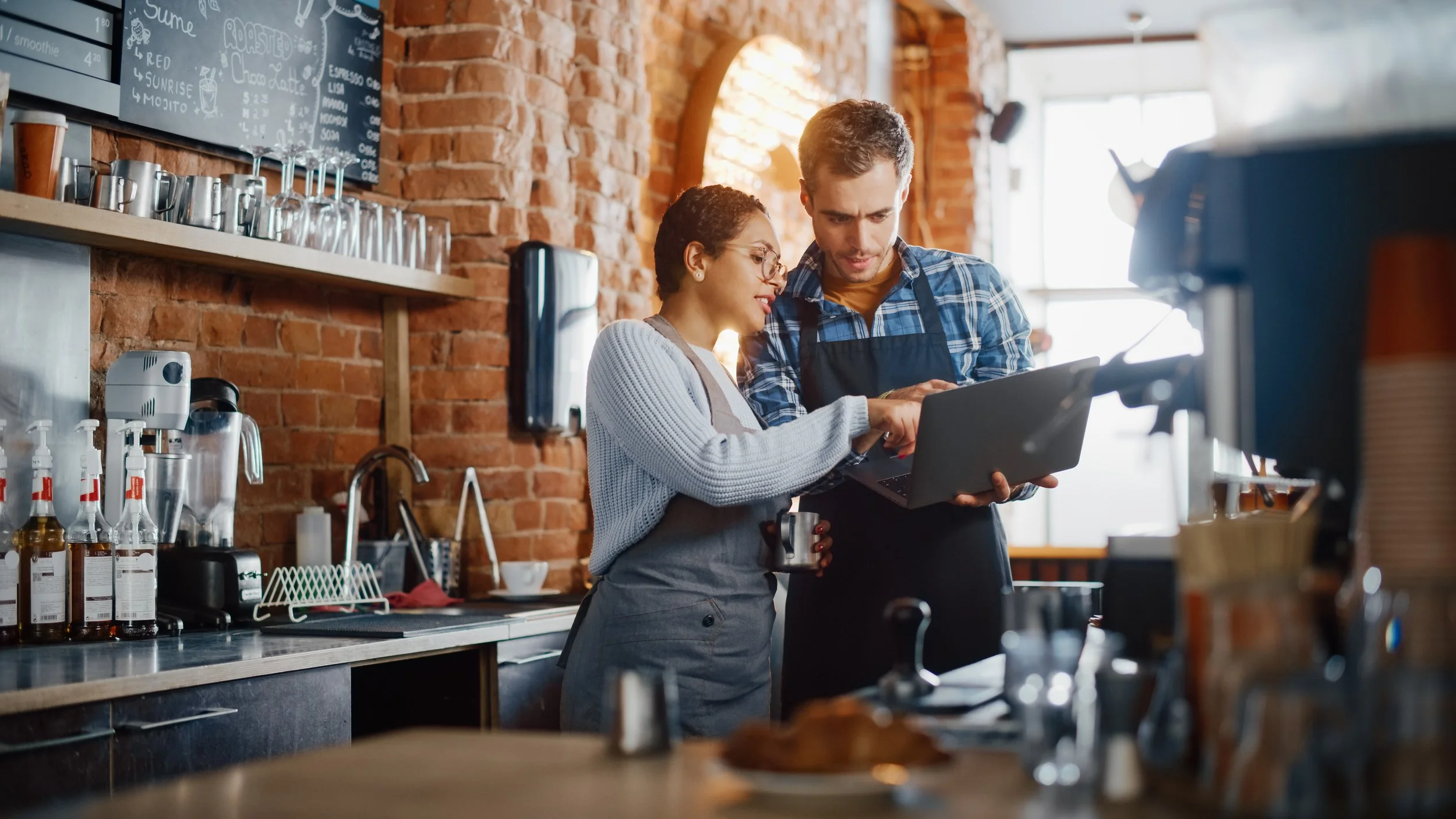 Two café owners behind the counter of their brick-walled coffee shop, looking at a laptop together.
