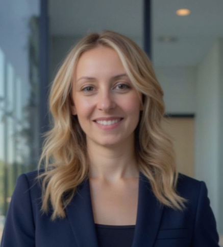 A young woman with blonde hair smiling, wearing a navy blazer, in an indoor office setting.
