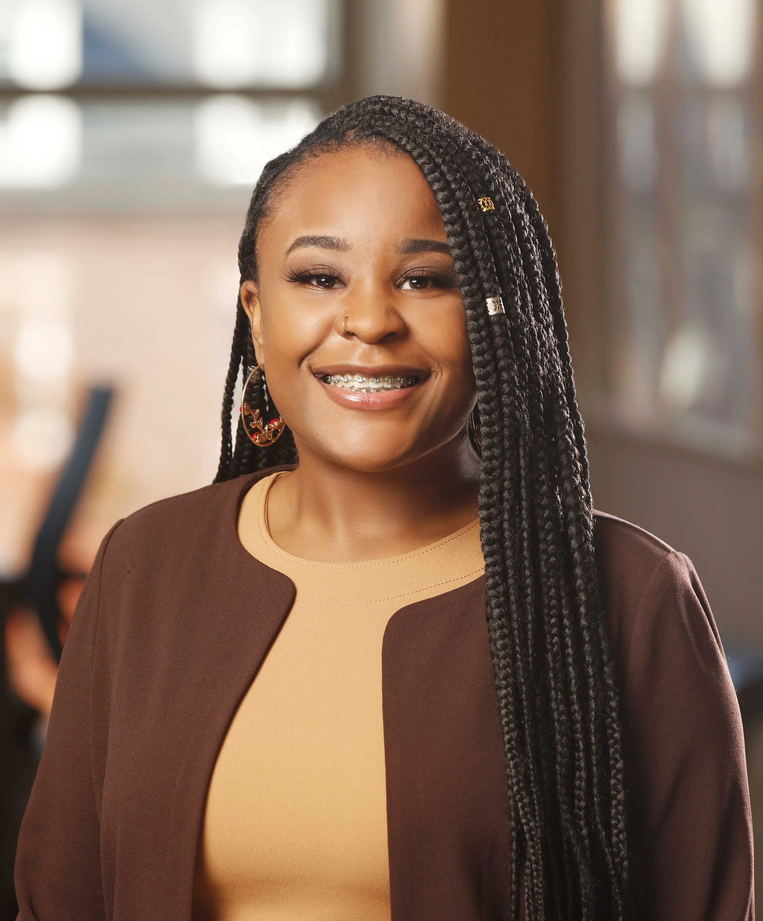 Close-up of a smiling young woman with braids, wearing braces, hoop earrings, and a nose ring, in a professional setting.