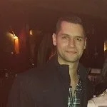 A man standing indoors, wearing a dark jacket and a plaid shirt, smiling at the camera.