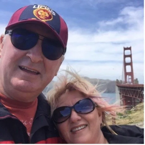 A smiling couple wearing sunglasses taking a selfie with the Golden Gate Bridge in the background during daytime.