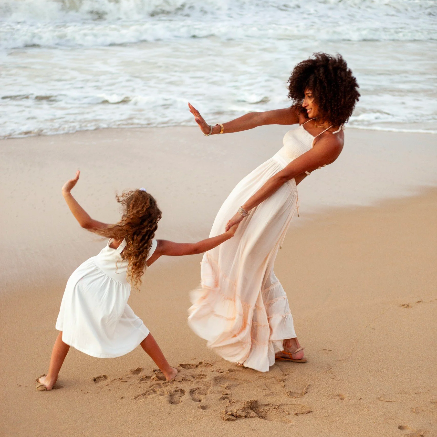 A woman and a girl dancing on the beach, holding hands, with ocean waves in the background.