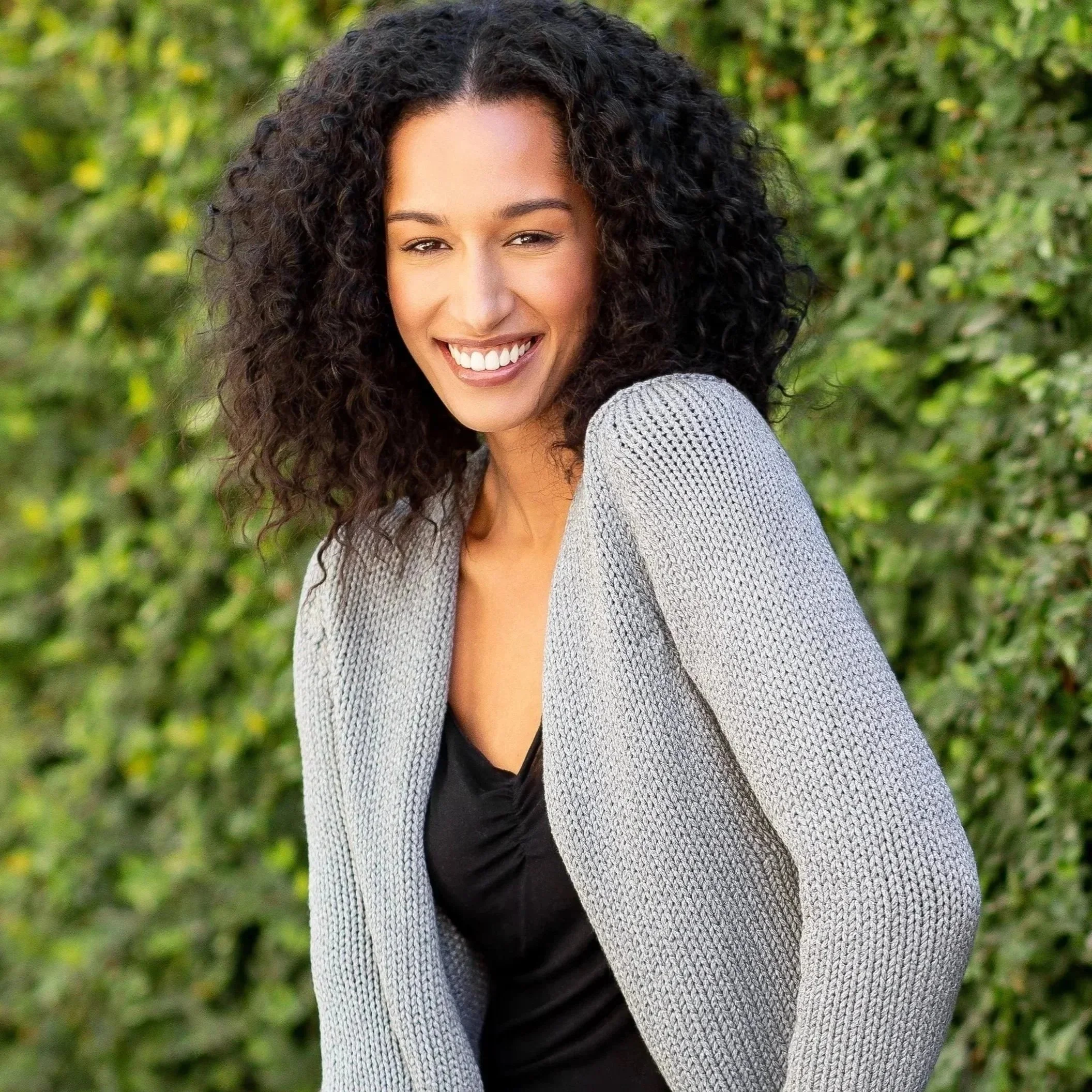 A woman with curly black hair smiling outdoors in front of green foliage, wearing a grey cardigan over a black top.