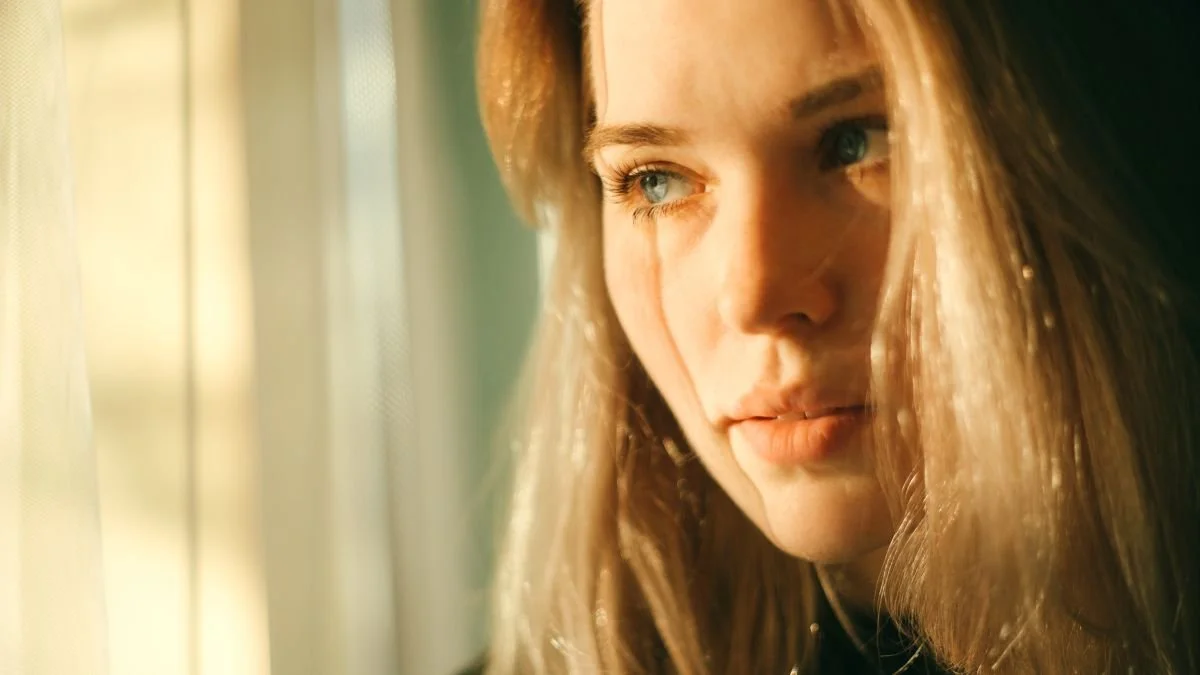 close up of a blonde women by a window. Signs your energy may be out of balance happen from chakra imbalance , nervous system overload, and energy imbalance. Energy medicine can provide a support system to help you return to balance.