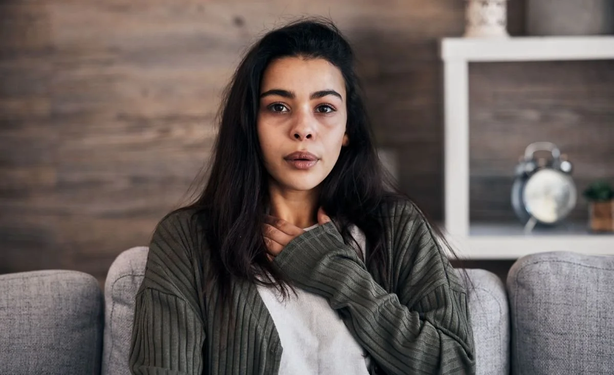 Dark haired woman, in living room, hand on her chest. Somatic awareness, emotional intelligence and other holistic wellness practices all balance the energy system on different levels of the aura.