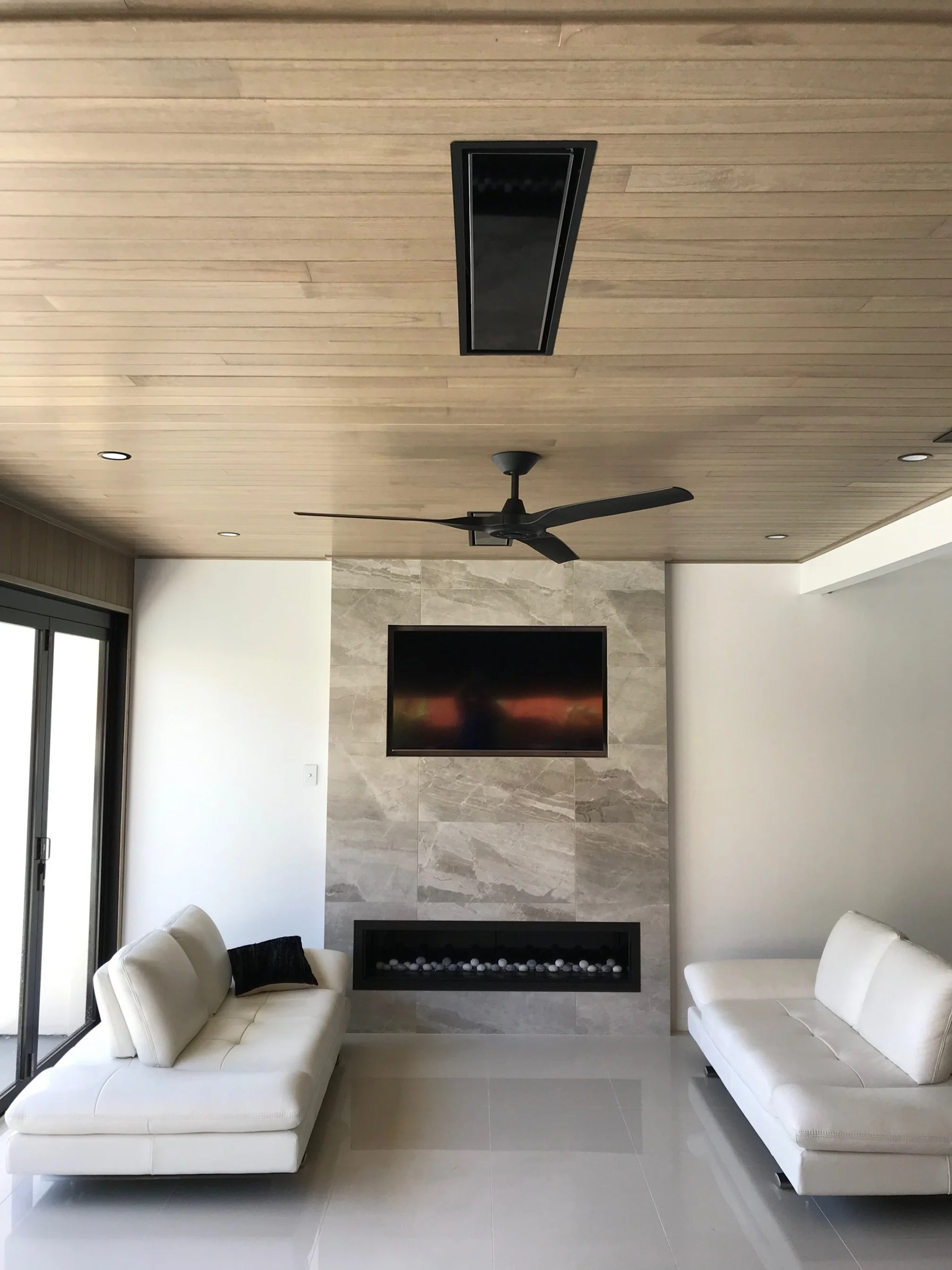 Modern indoor/outdoor living room with white sofas, wall-mounted TV, electric fireplace, ceiling fan, and light wood cladded ceiling.