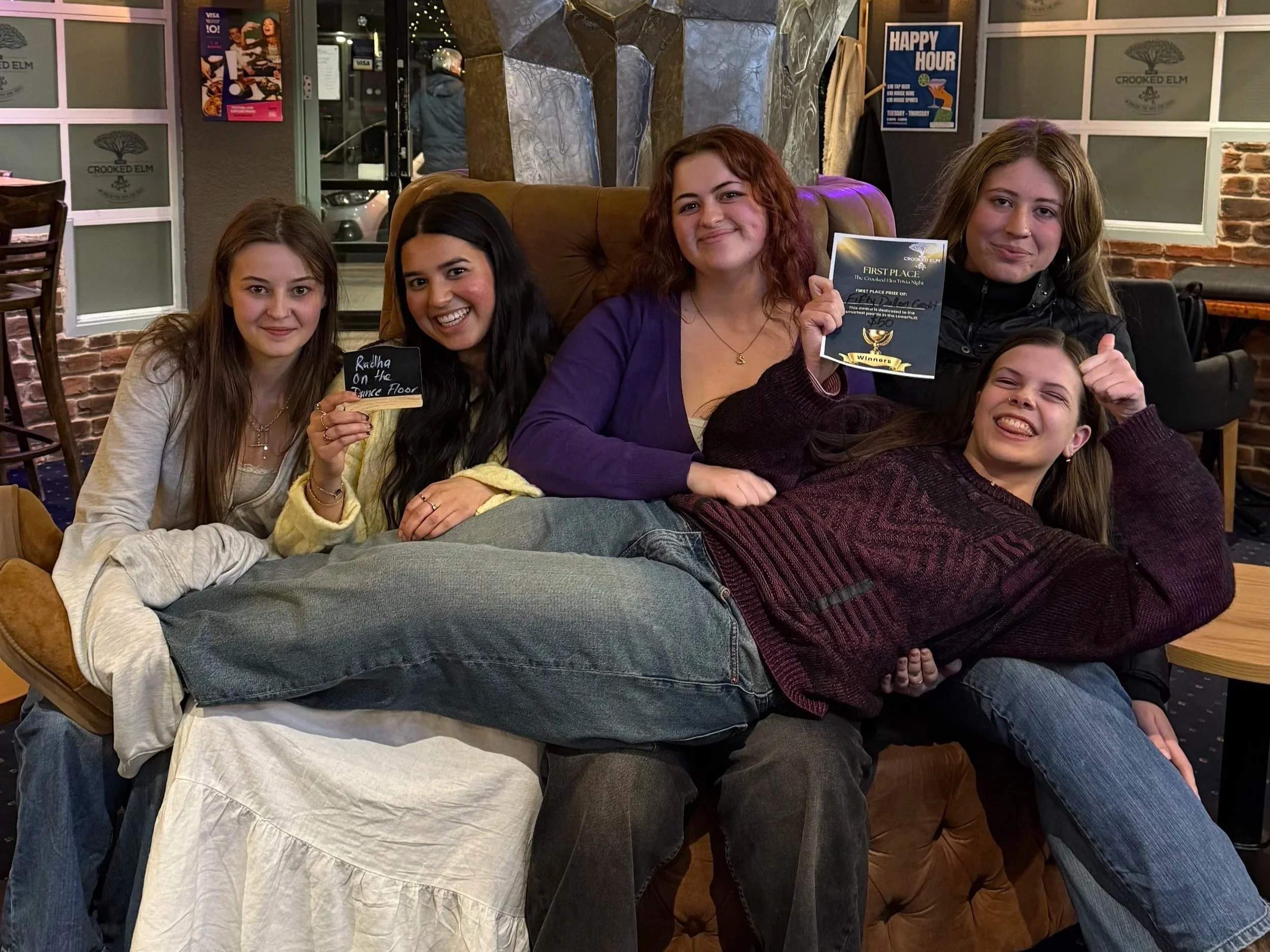 Group of five young women sitting on a couch in a restaurant or cafe, celebrating a dance competition win. Two women are holding awards, and one woman has a small chalkboard sign that says 'Racha on the Dance Floor.' They are smiling and making celebratory gestures, with a cozy, decorated background.